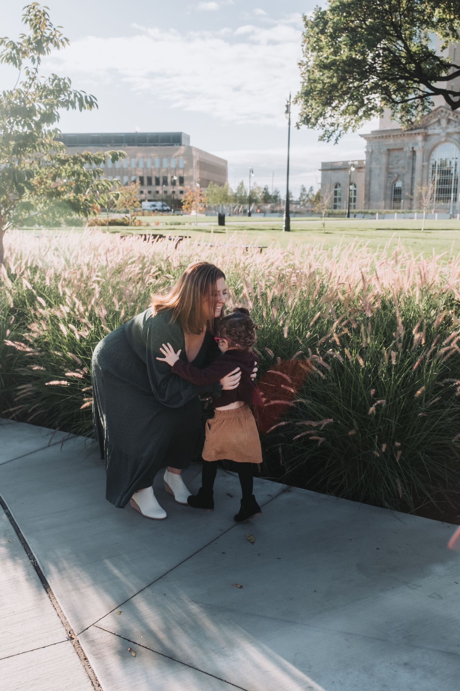 A woman crouches down to hug a young child near ornamental grasses in a sunny urban park setting.