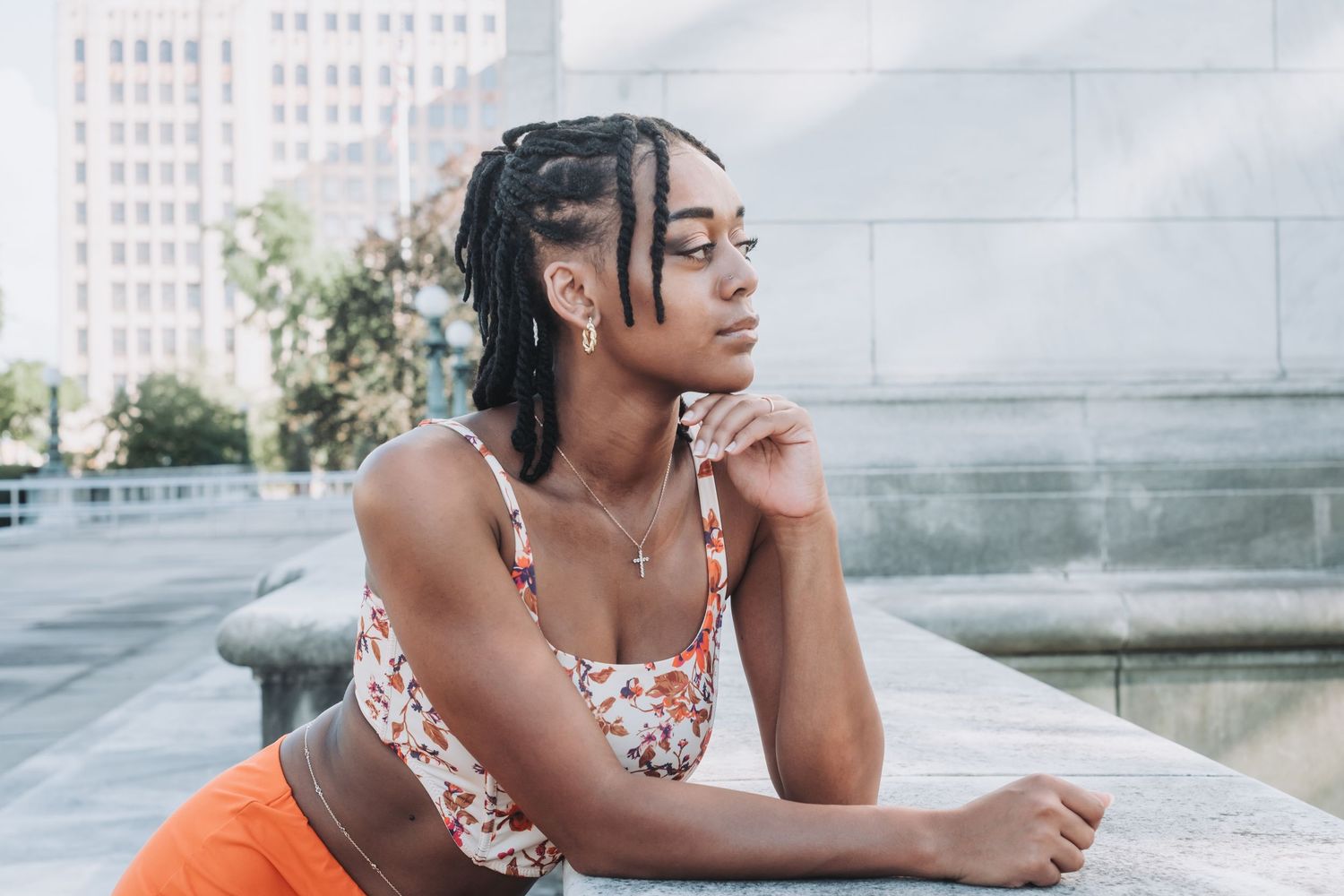 Young Black woman with braids wearing floral crop top and orange skirt, posing outdoors near urban fountain.