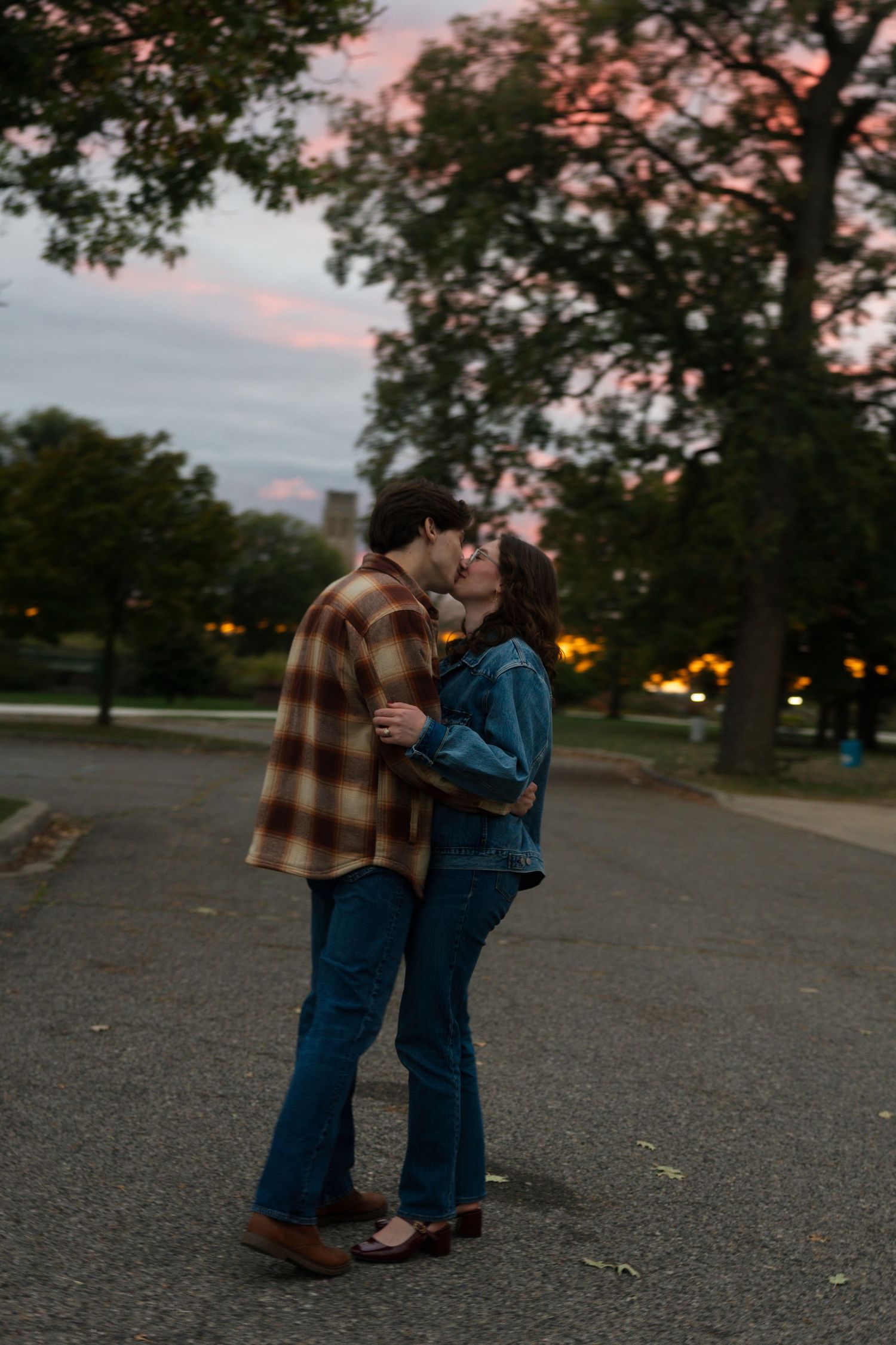 Couple sharing a romantic kiss at dusk in a park, surrounded by trees with a pink sunset sky backdrop.