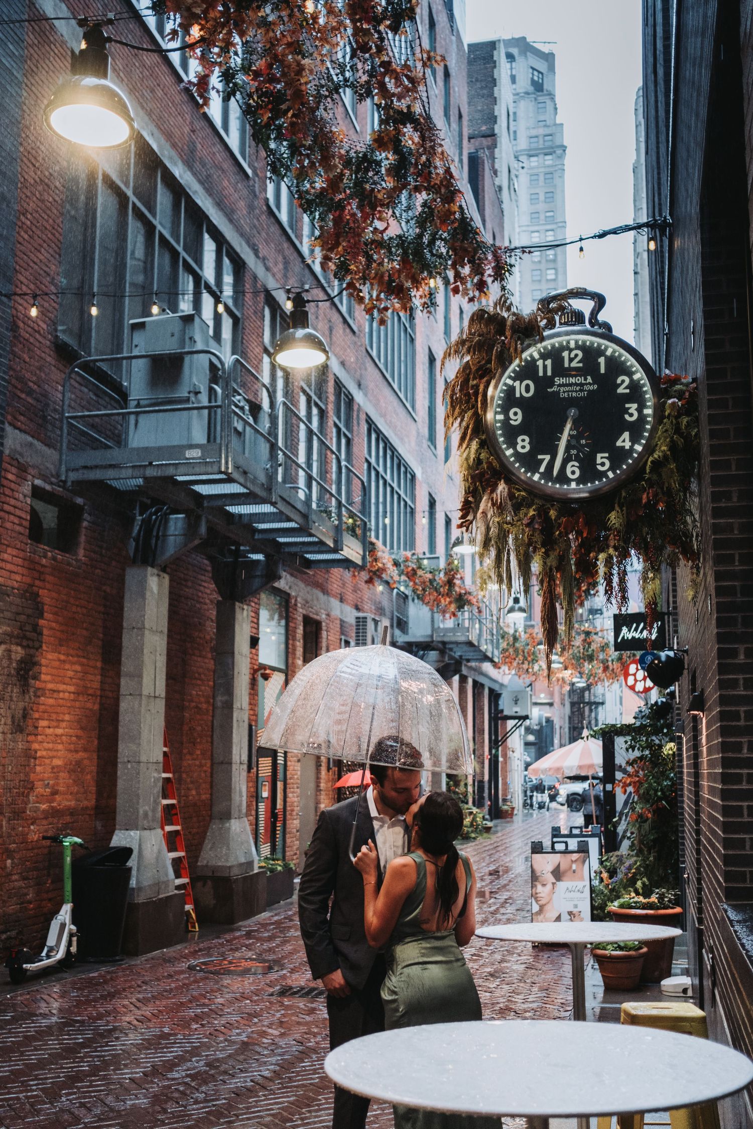 Couple kissing under clear umbrella in rainy brick alley with vintage clock and hanging florals.