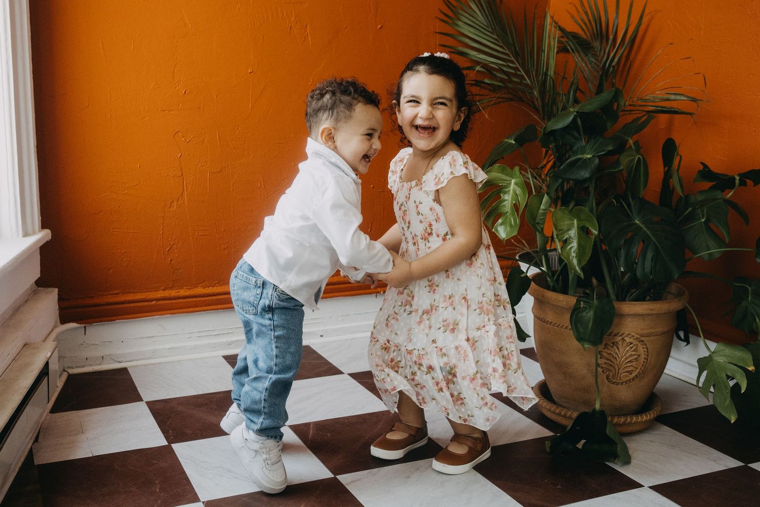 Two happy children laughing and holding hands on a checkered floor near a potted palm plant against an orange wall.