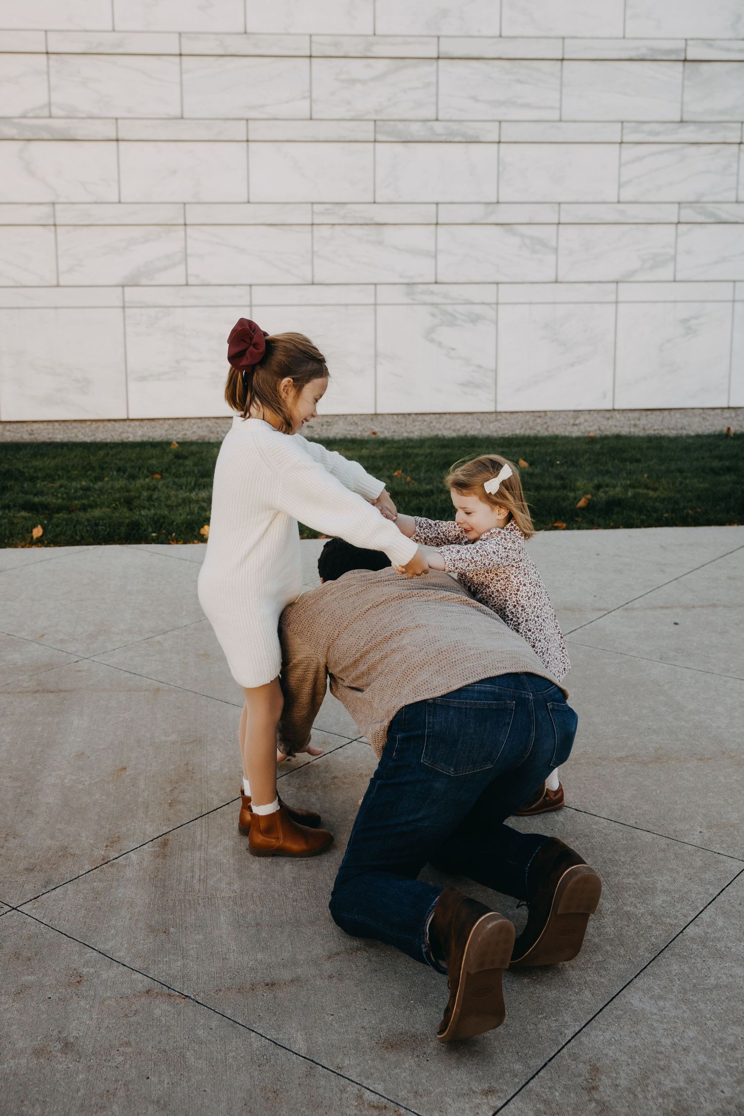 Two girls playfully interact with a kneeling adult outdoors near a marble wall on a tiled plaza.