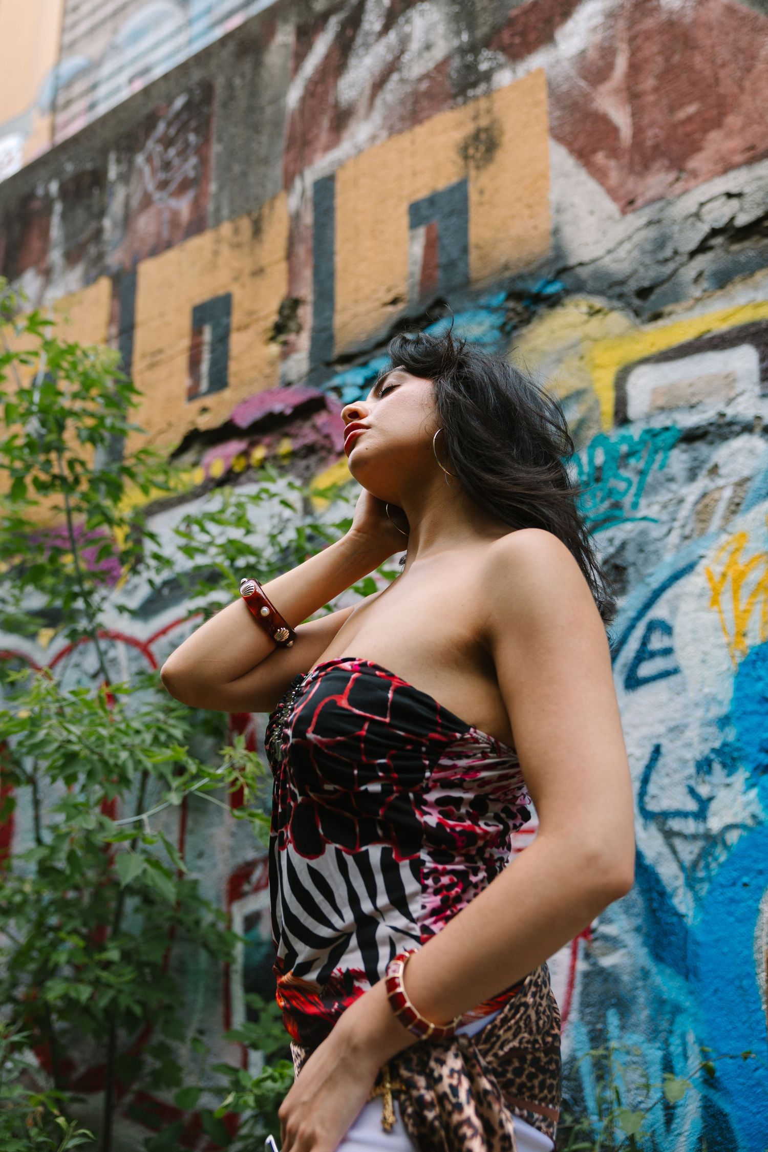 Young woman in strapless patterned dress poses confidently against a colorful graffiti wall outdoors.