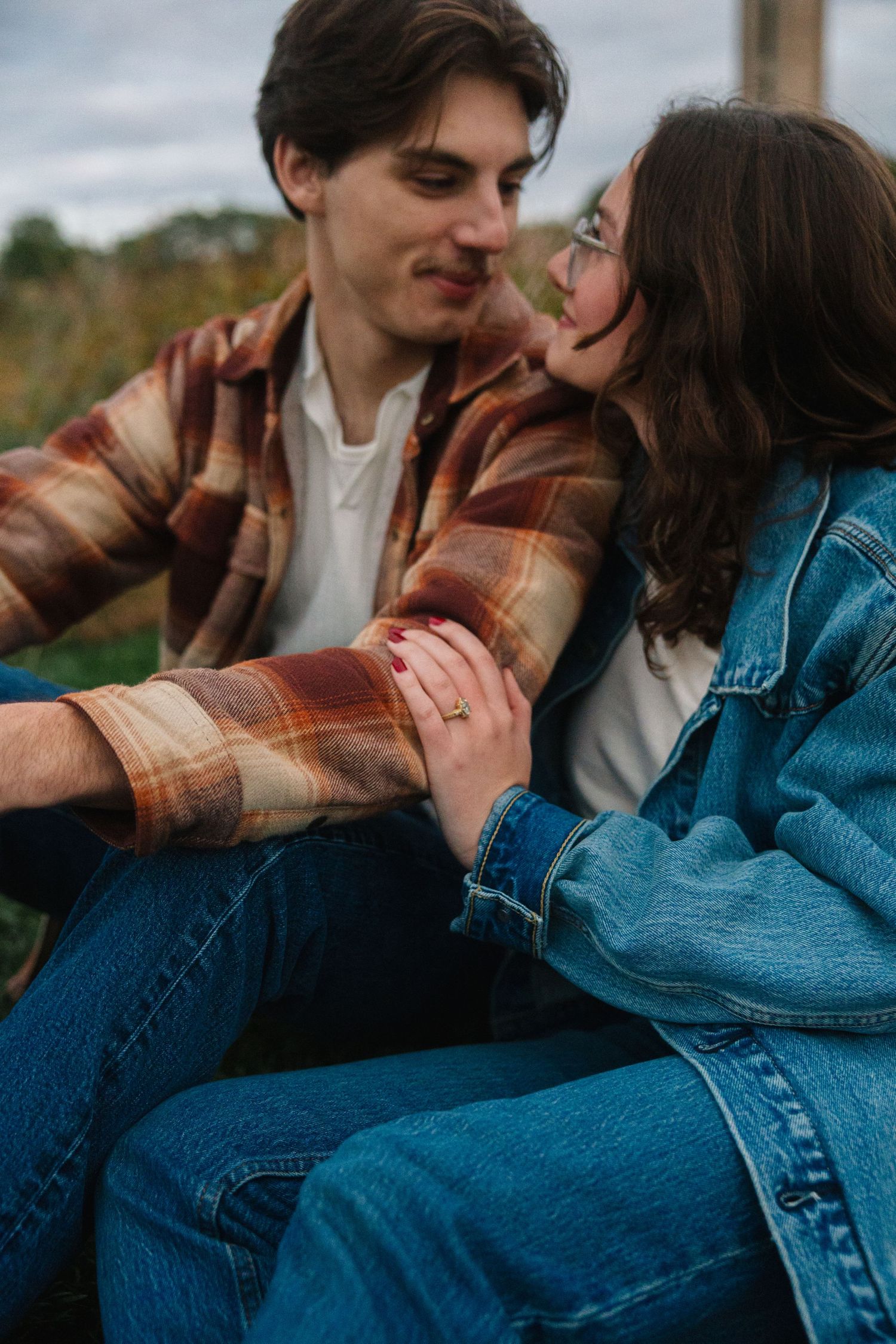 A couple sits closely outdoors, holding hands and smiling, dressed in plaid and denim jackets on a cloudy day.