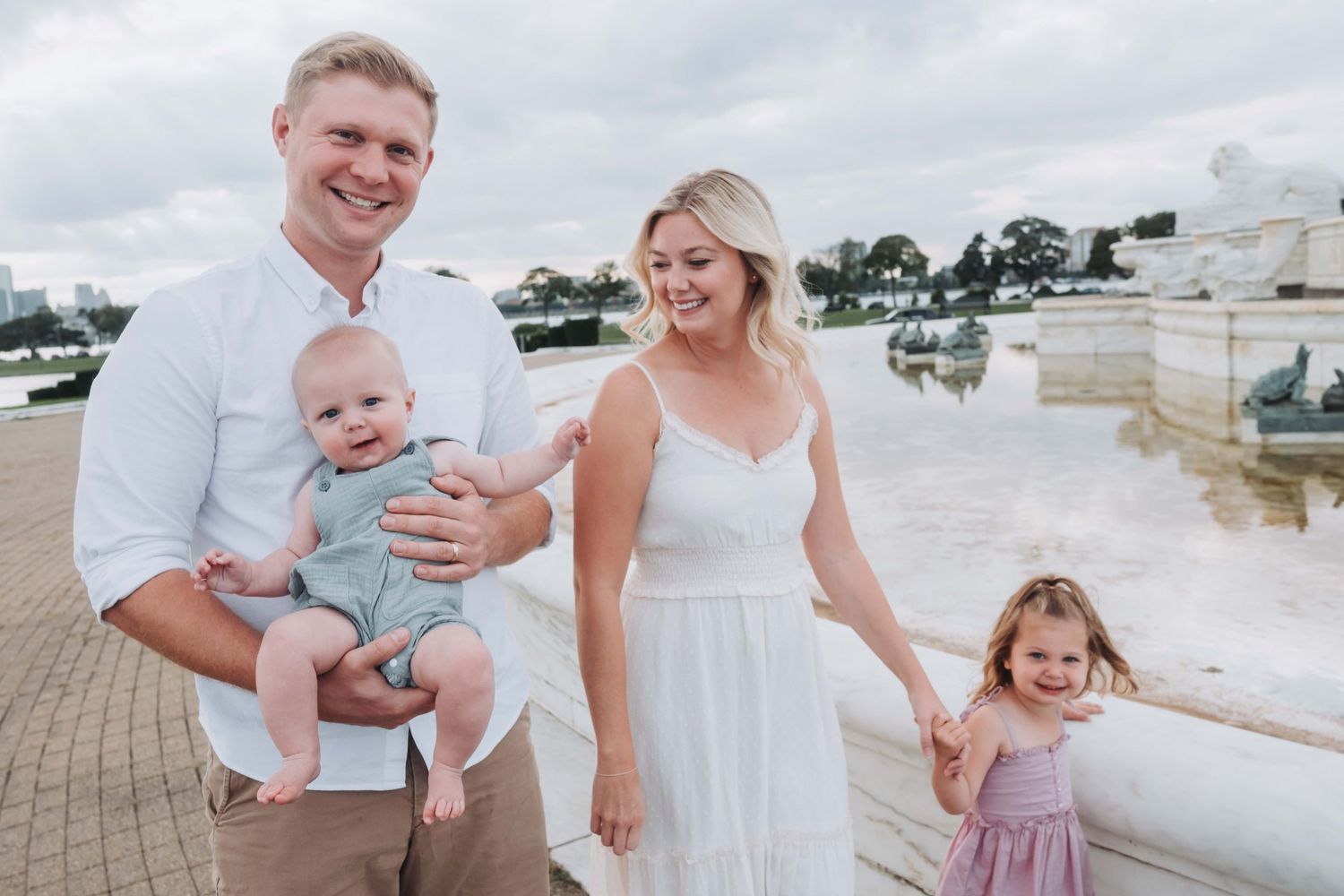 Happy family of four posing outdoors near a fountain, parents holding young children, dressed in white and pastel outfits.