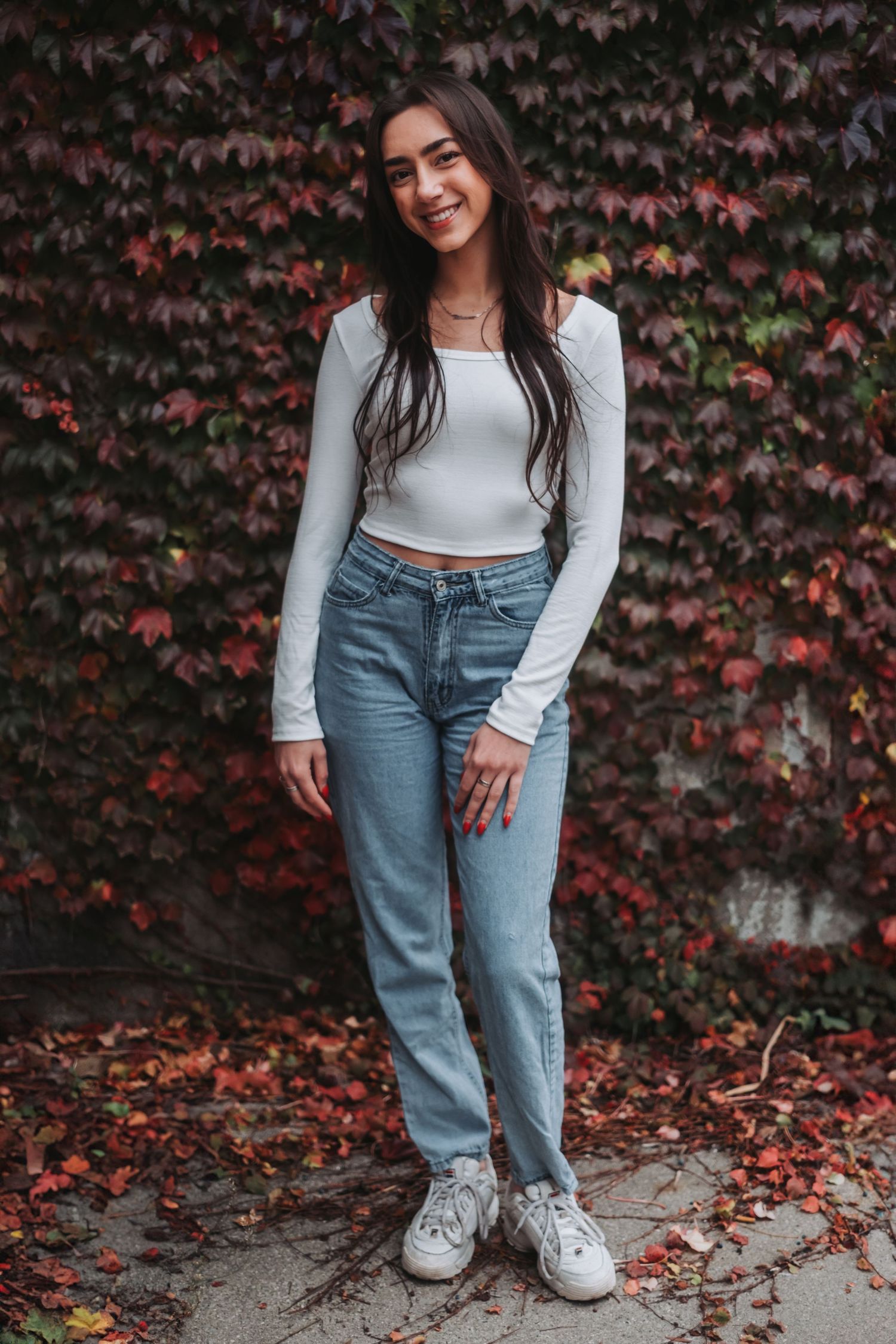 Young woman in white crop top and blue jeans smiling outdoors against a red autumn ivy wall.
