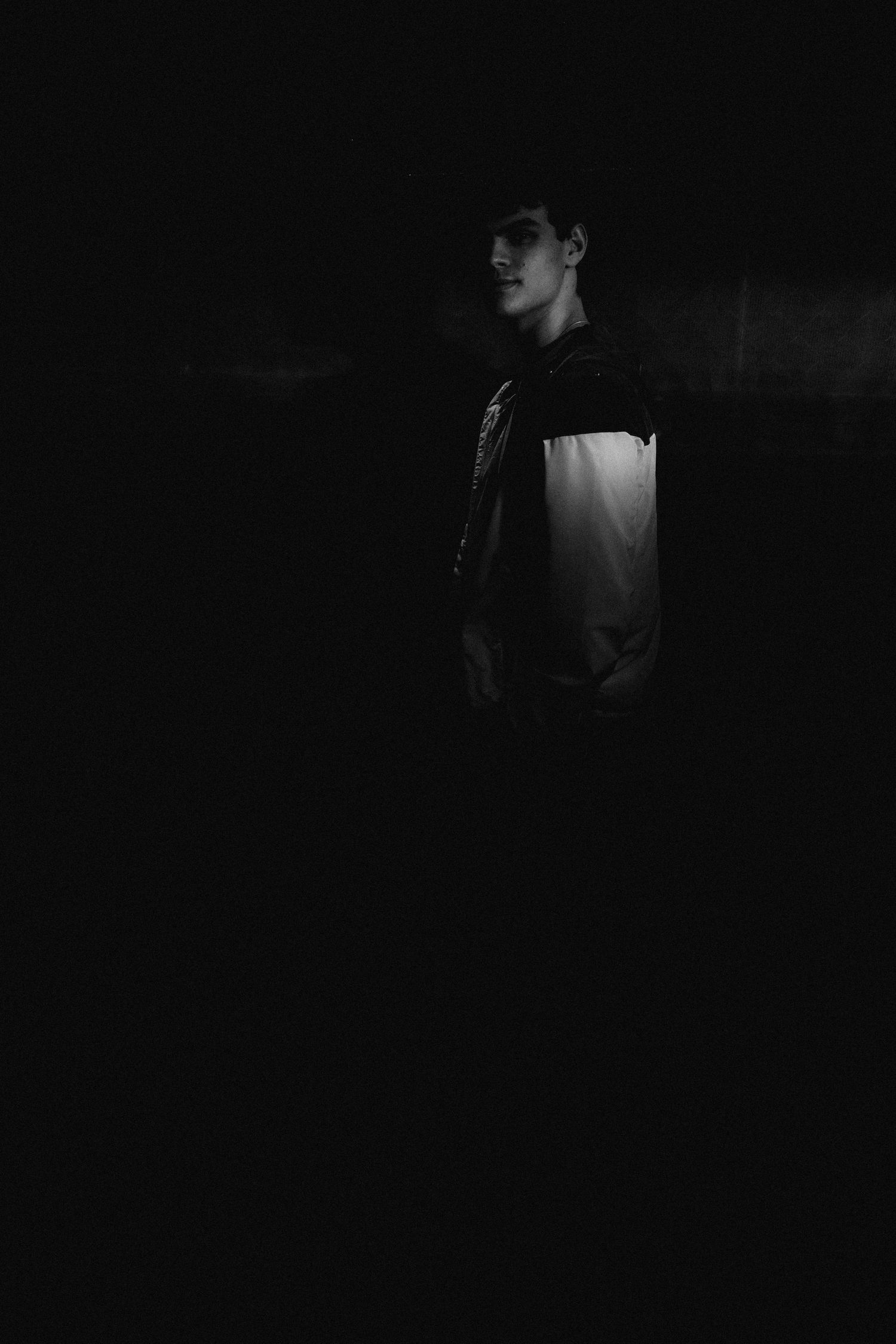 Black and white portrait of a young man standing in dramatic dark shadow lighting wearing a jacket.
