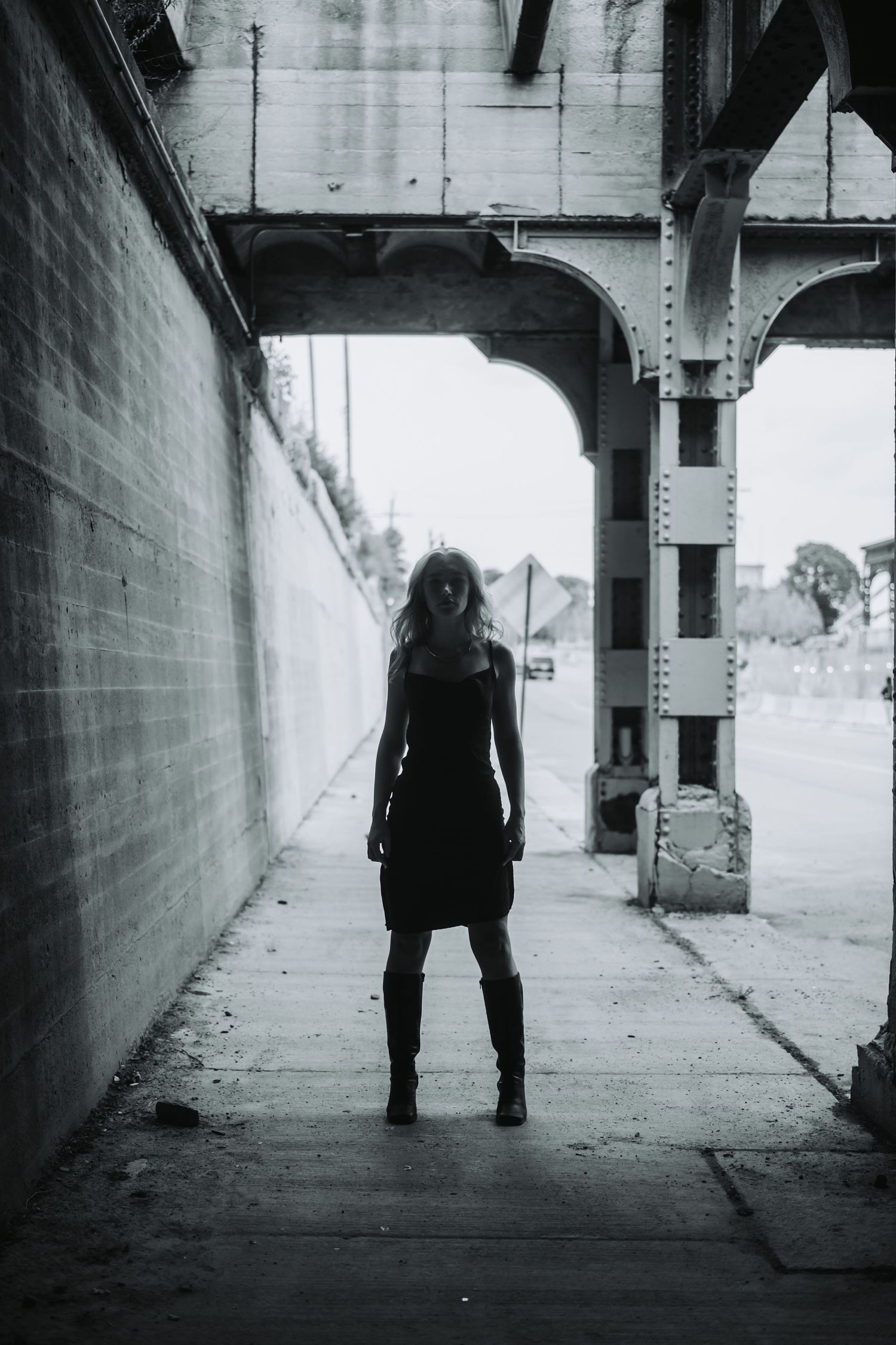 Woman in black dress and boots standing under urban bridge overpass in dramatic black and white photography.