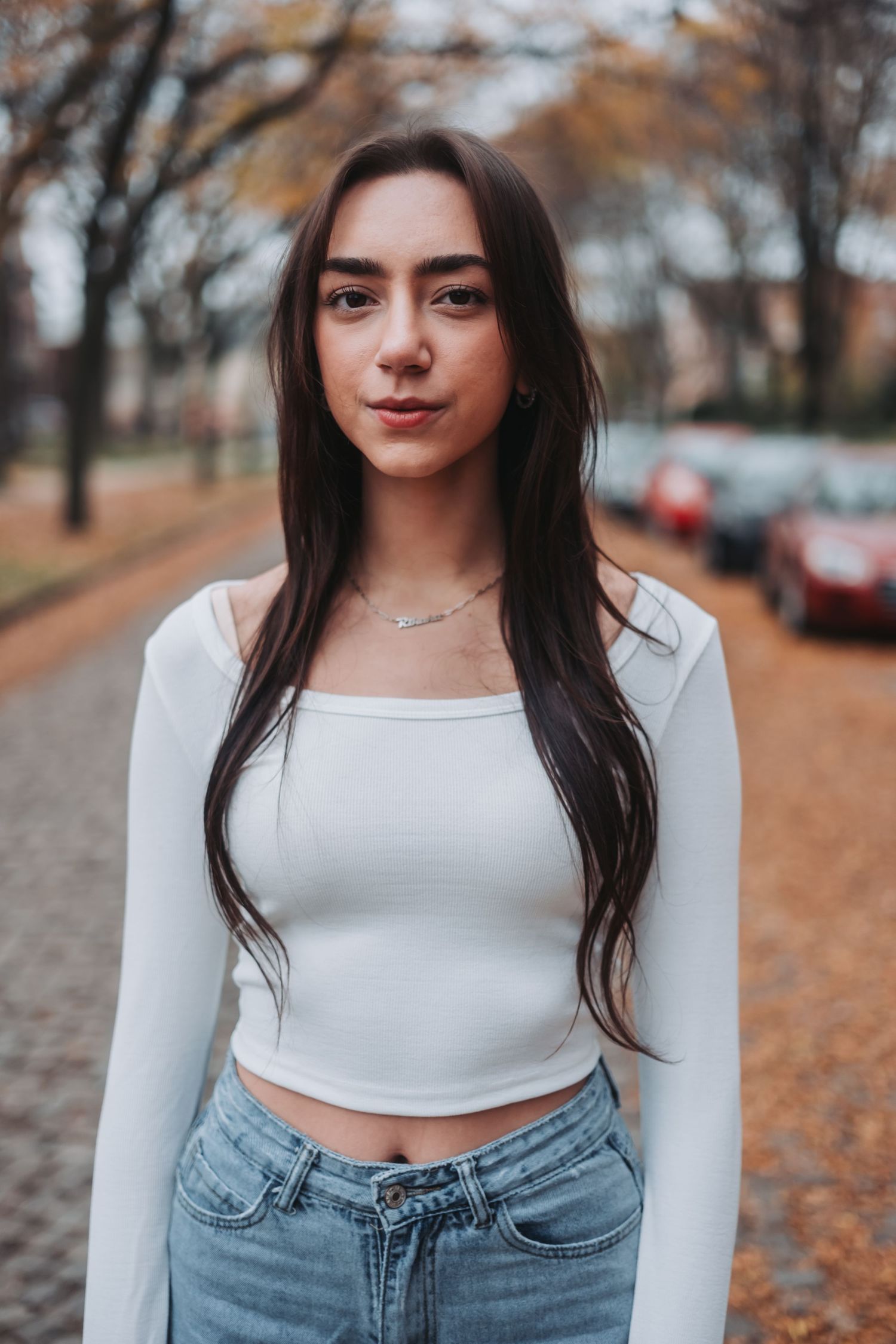 Young woman with long dark hair wearing a white crop top and jeans standing on a tree-lined street in autumn.