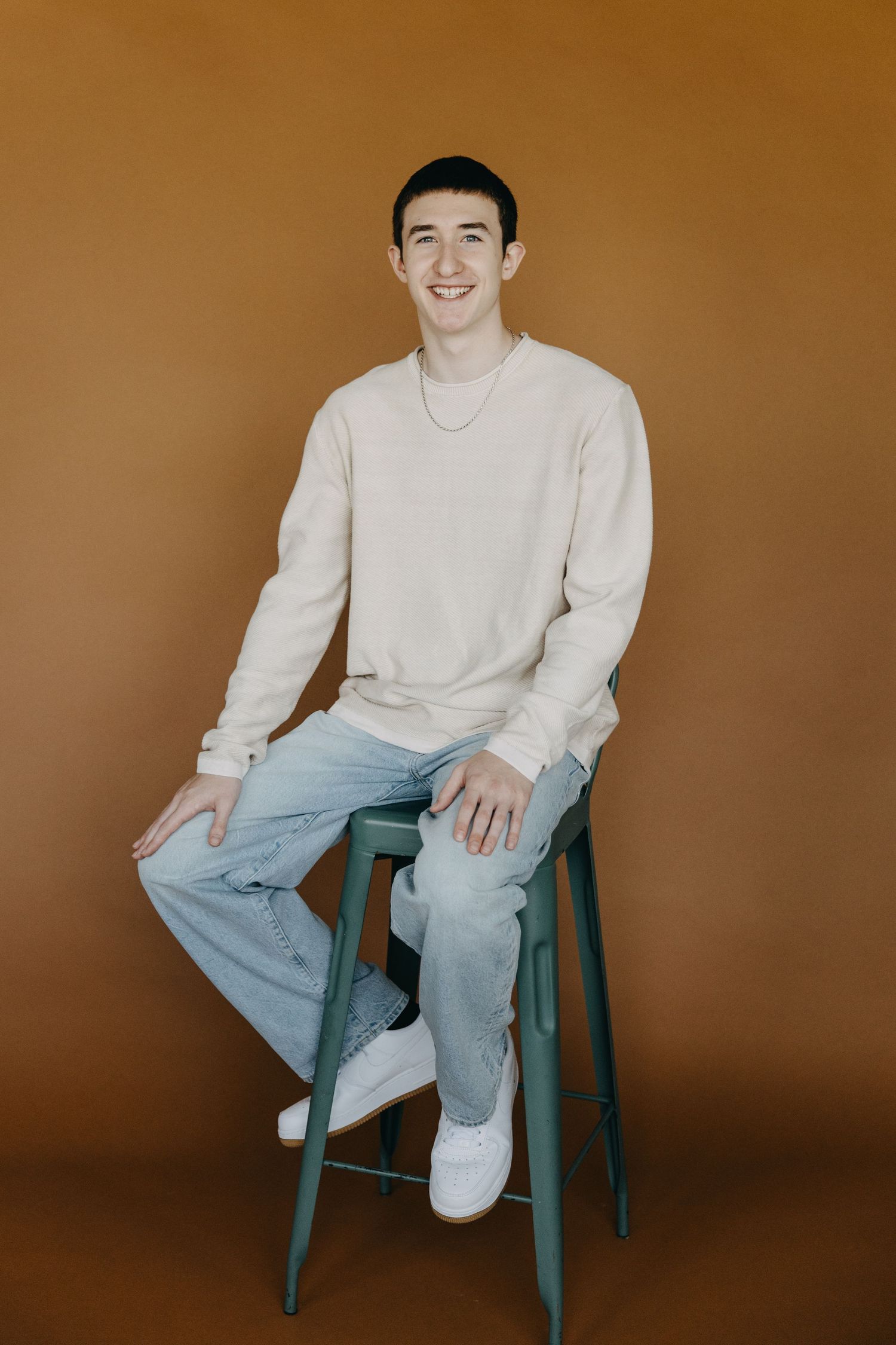 Young man smiling, seated on teal stool, wearing beige sweatshirt and light jeans, against warm brown background.