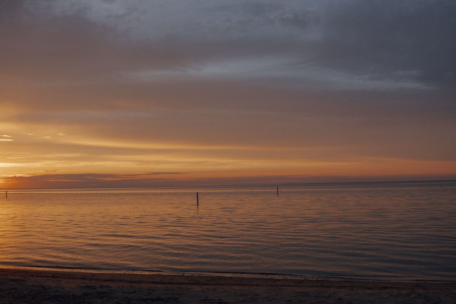 Serene beach sunset with golden orange sky reflecting on calm ocean waters, with distant marker buoys on the horizon.