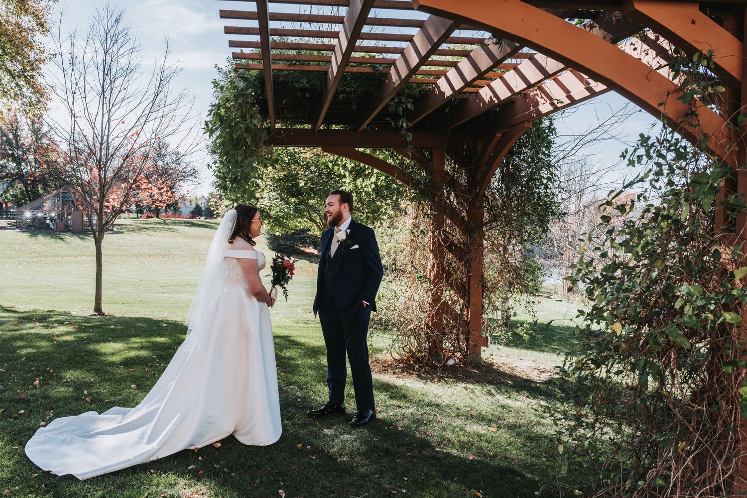 Bride and groom share first look moment under rustic wooden pergola surrounded by autumn foliage.