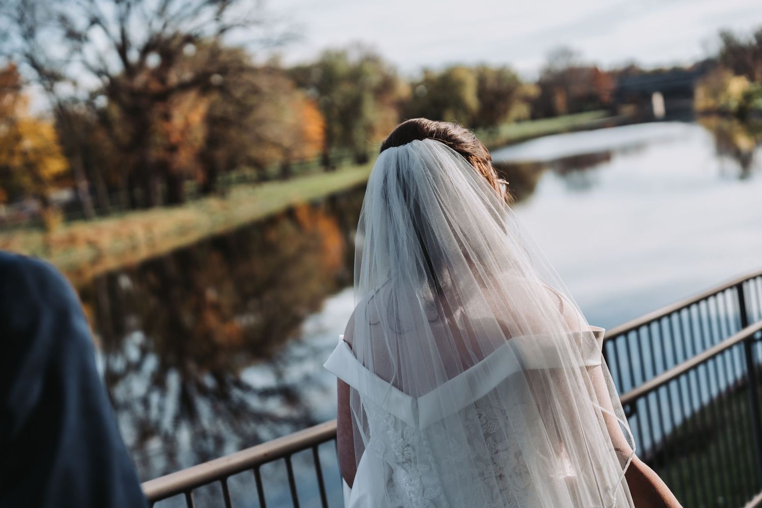 Bride in white gown and veil standing on bridge overlooking scenic autumn river landscape.