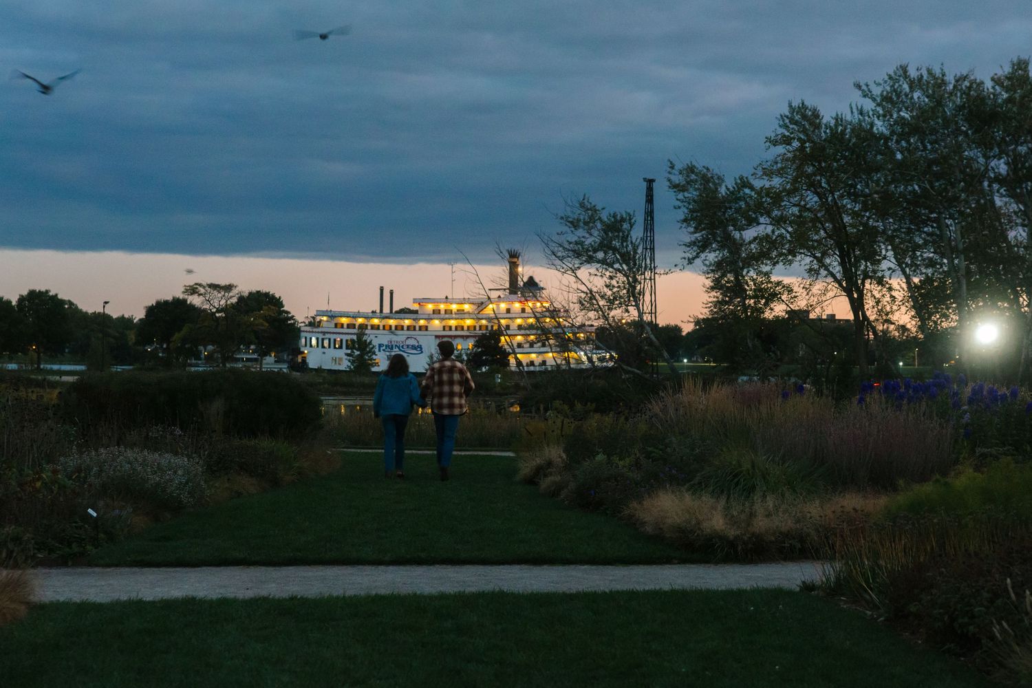 Couple walking through a scenic park at dusk with a riverboat and glowing lights reflecting on the water.
