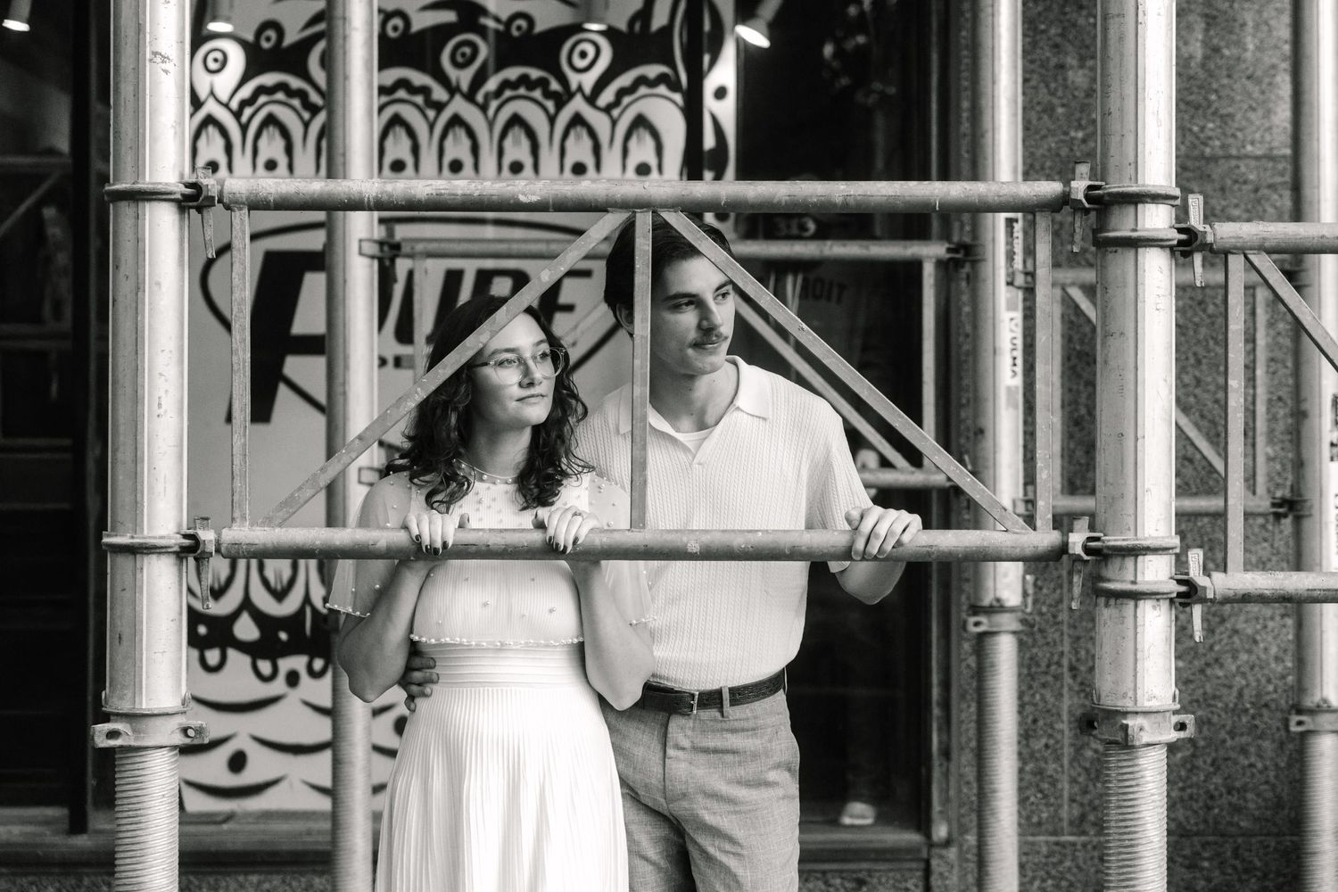 Black and white photo of a couple standing together among scaffolding poles outside a building with decorative windows.