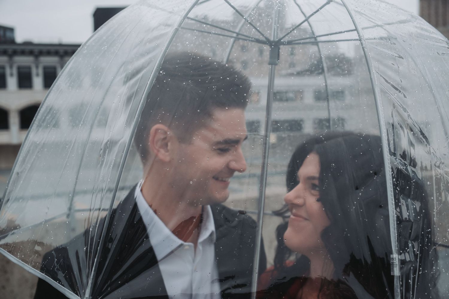 Couple smiling at each other under a clear bubble umbrella in the rain with a city building in the background.