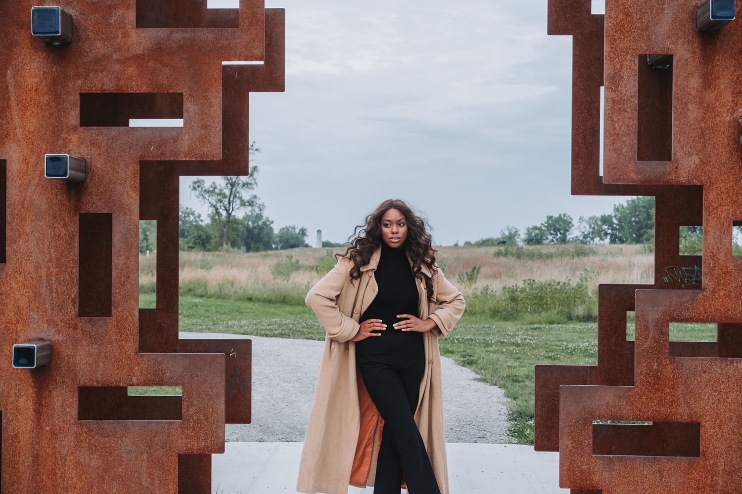 Woman in beige trench coat and black outfit posing between rustic geometric metal sculpture frames outdoors.