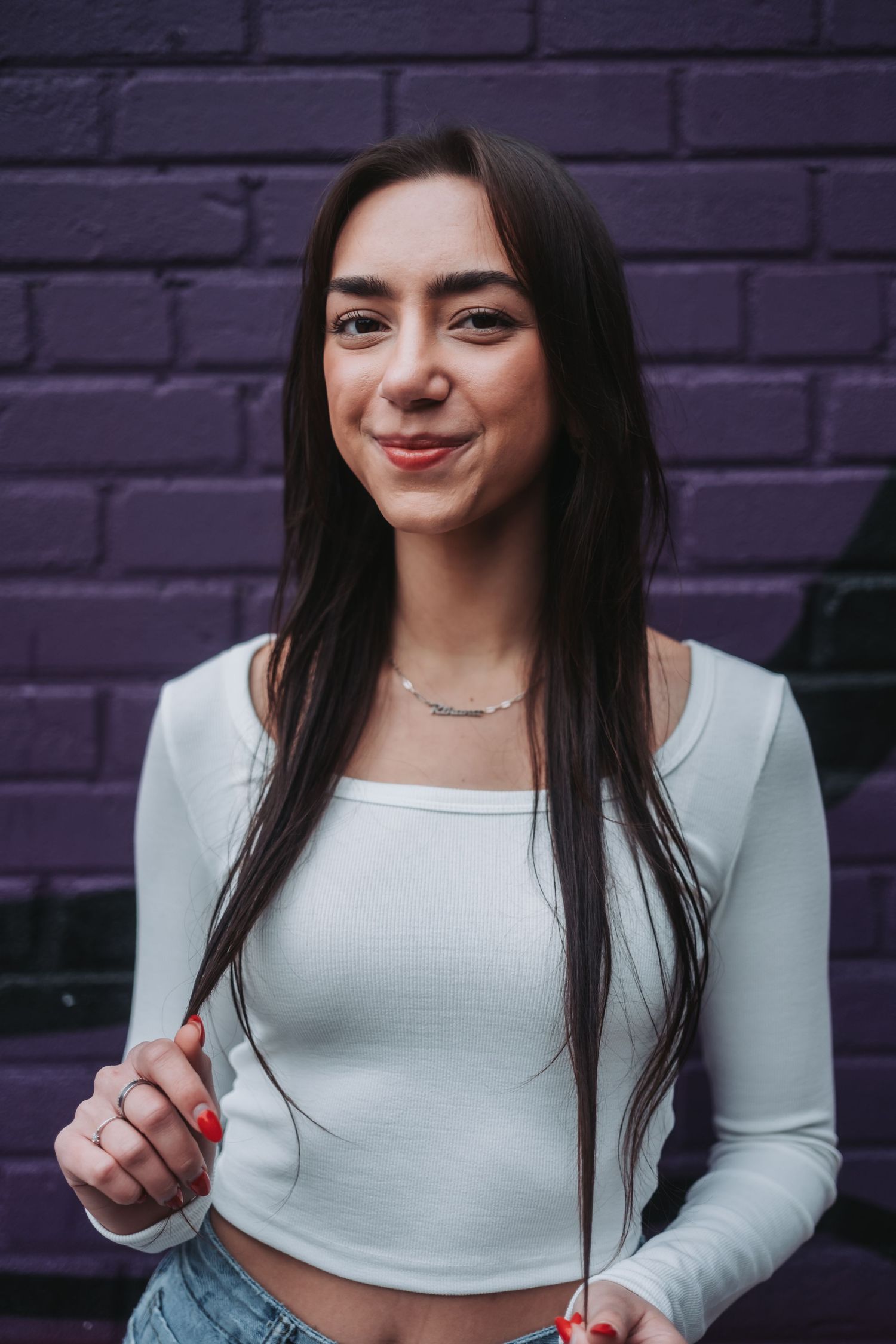 A woman in a white crop top smiles against a purple brick wall background.