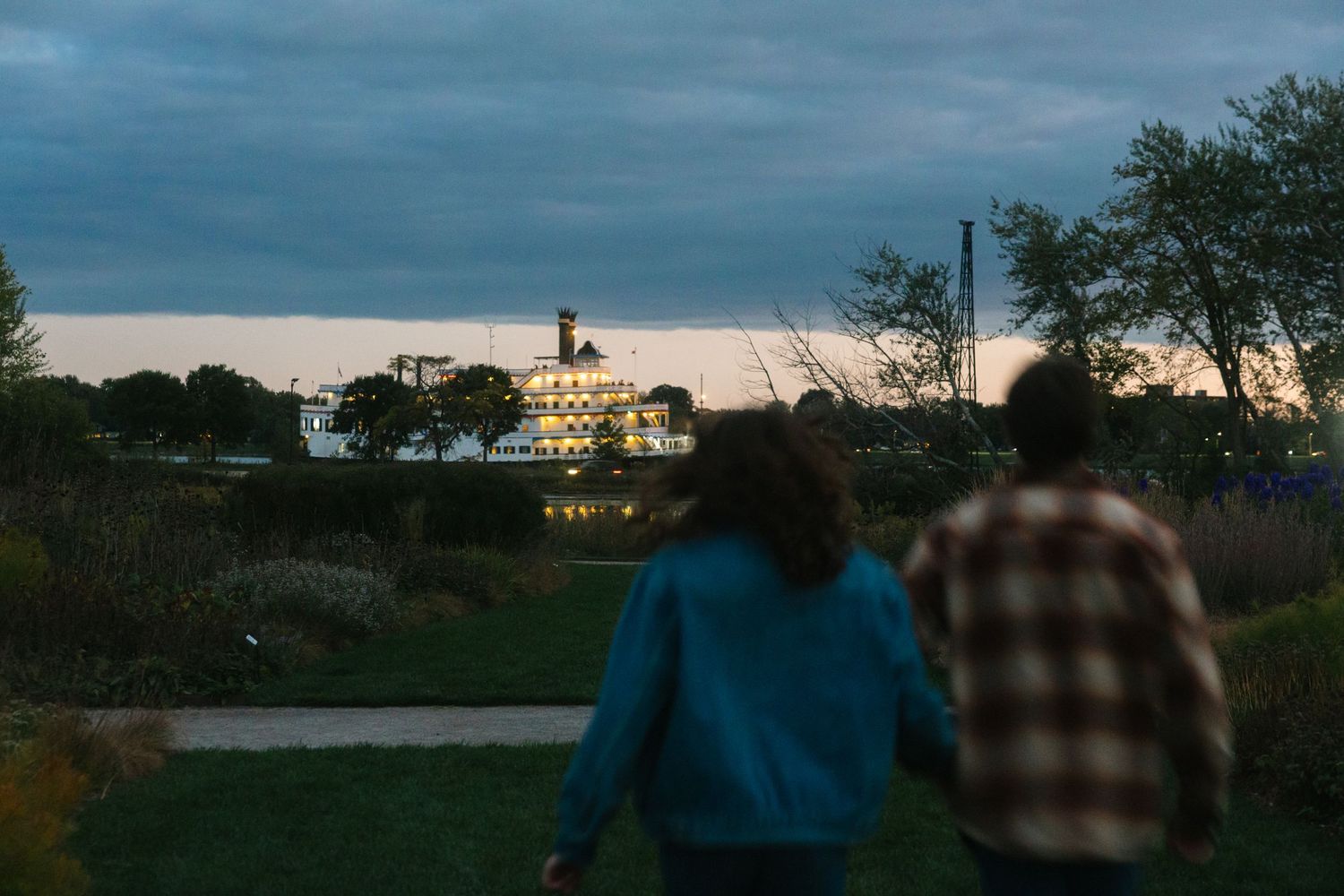 Couple walking hand in hand through a park at dusk with illuminated building in background.