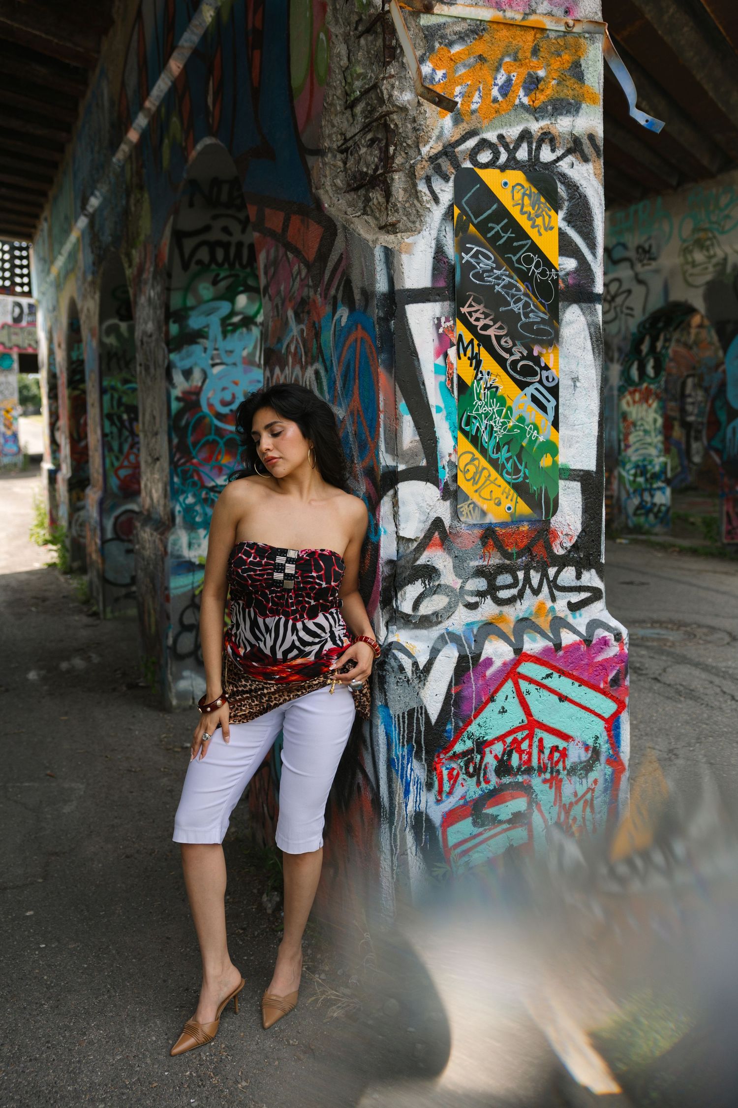 Young woman in strapless top and white capris posing against colorful graffiti-covered urban wall.