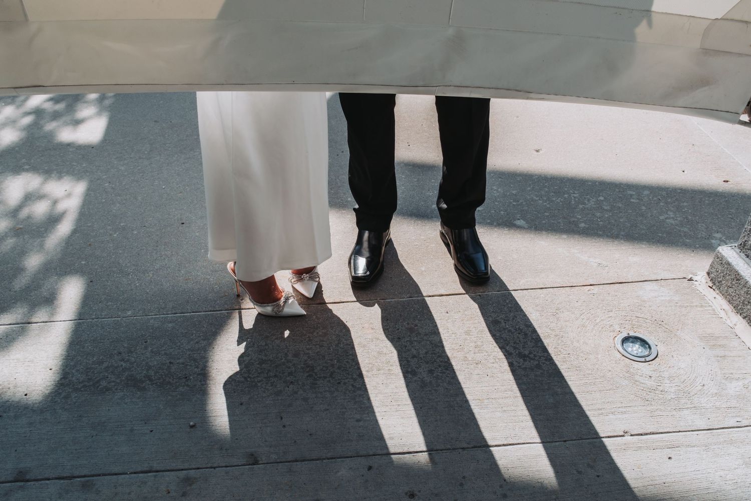 Close-up of bride in white gown with silver heels and groom in black suit with dress shoes on a sunlit sidewalk.