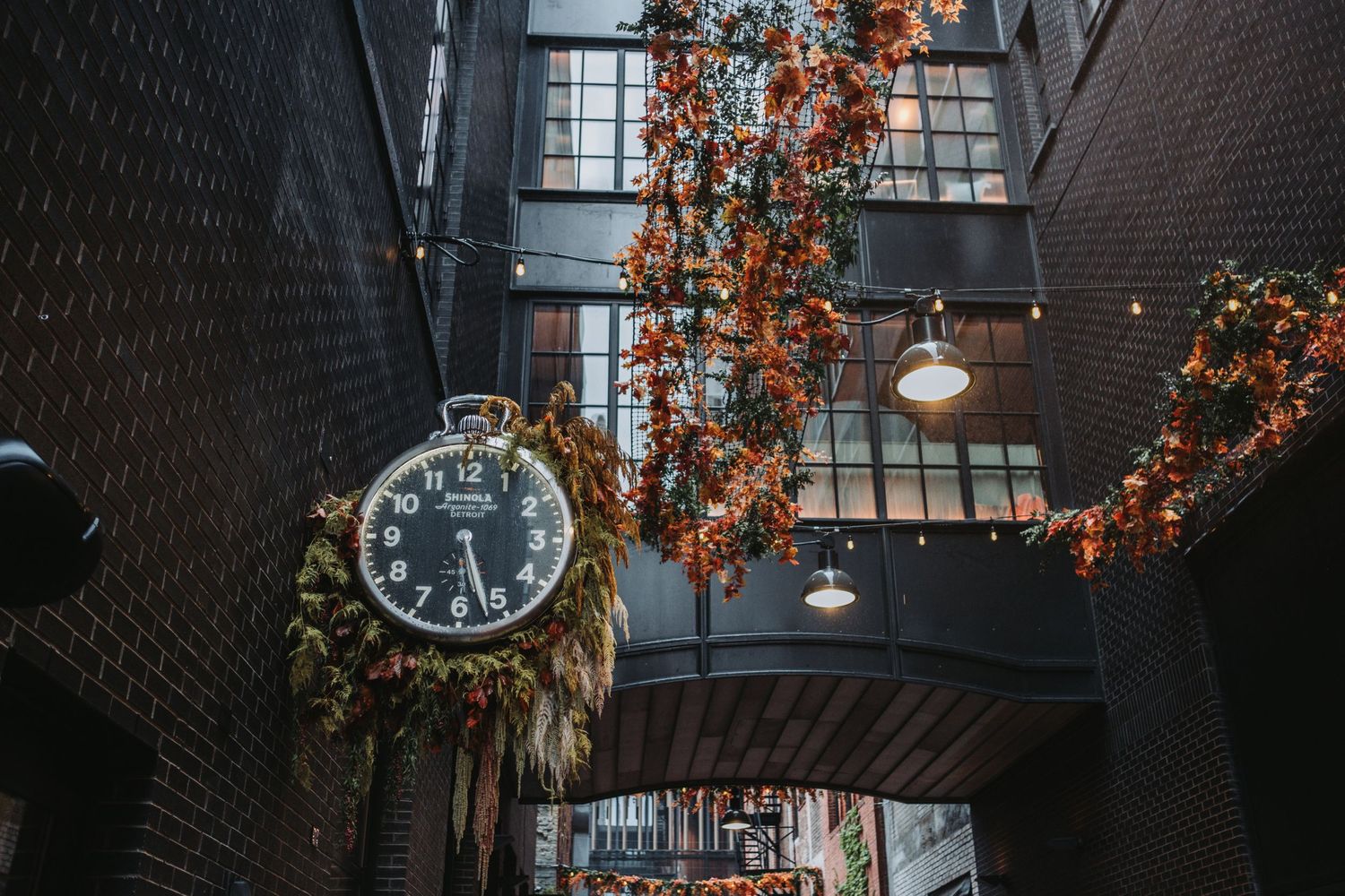 Vintage clock adorned with greenery hangs in a brick alley with festive floral garlands and warm pendant lights.