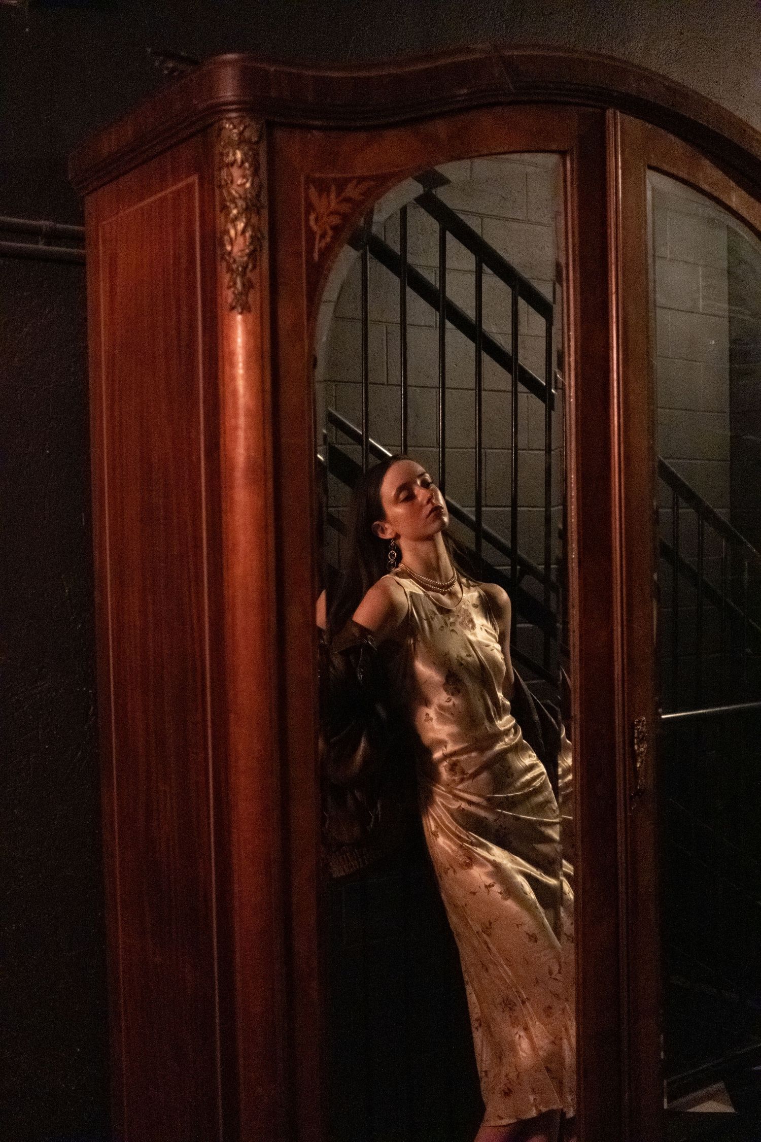 Woman in golden satin gown poses elegantly inside ornate wooden wardrobe with arched mirror, dramatic moody lighting.