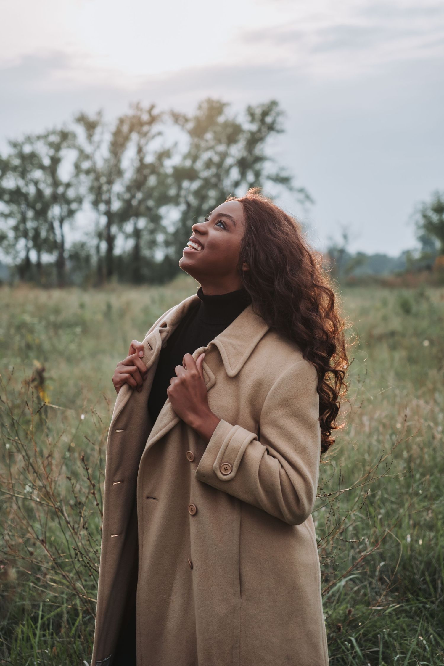 A smiling woman in a beige trench coat stands in a green field, looking up joyfully on a cloudy day.