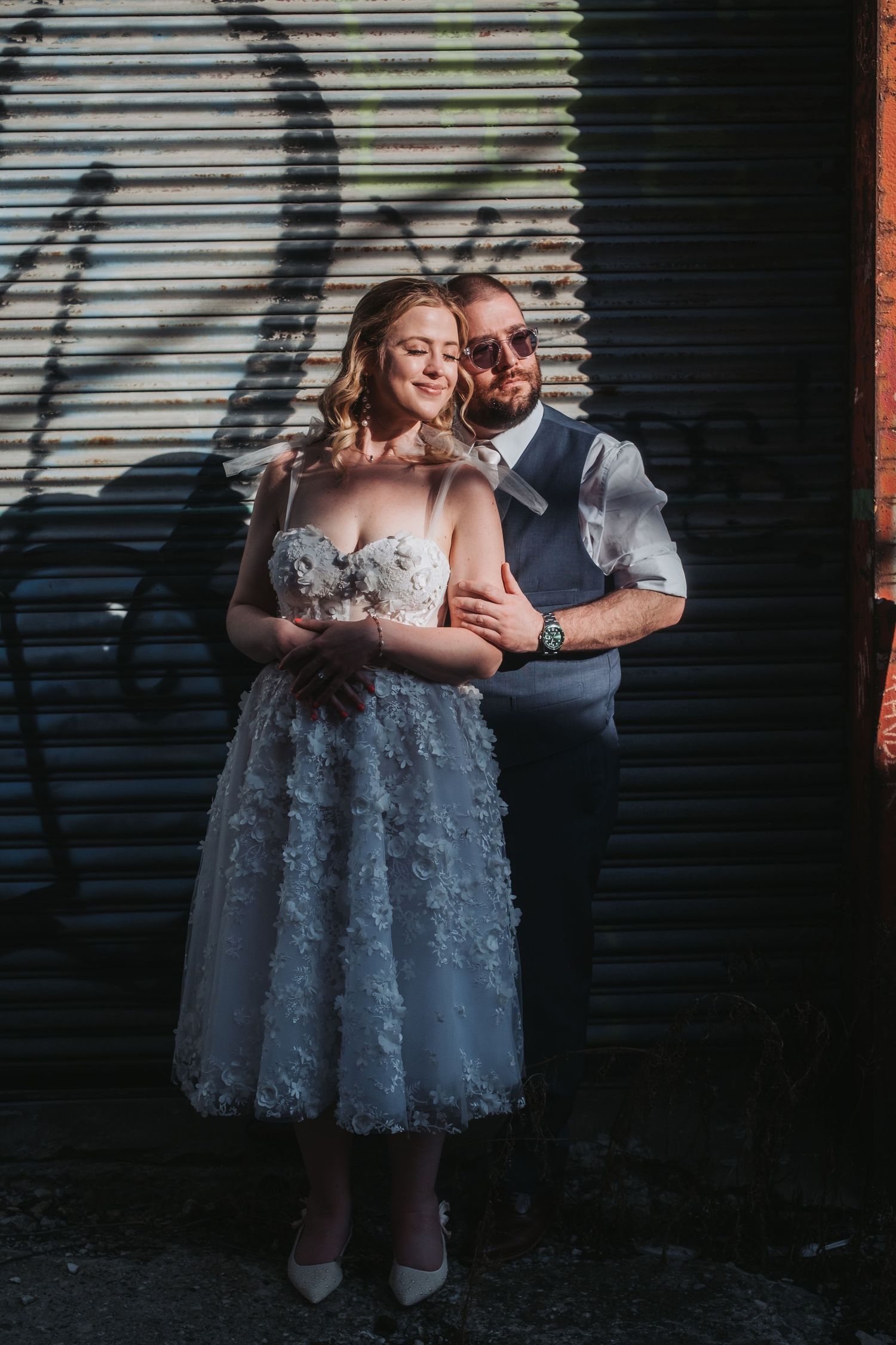 A bride in a floral white dress holds a bouquet as a groom in a vest embraces her against a graffiti wall.