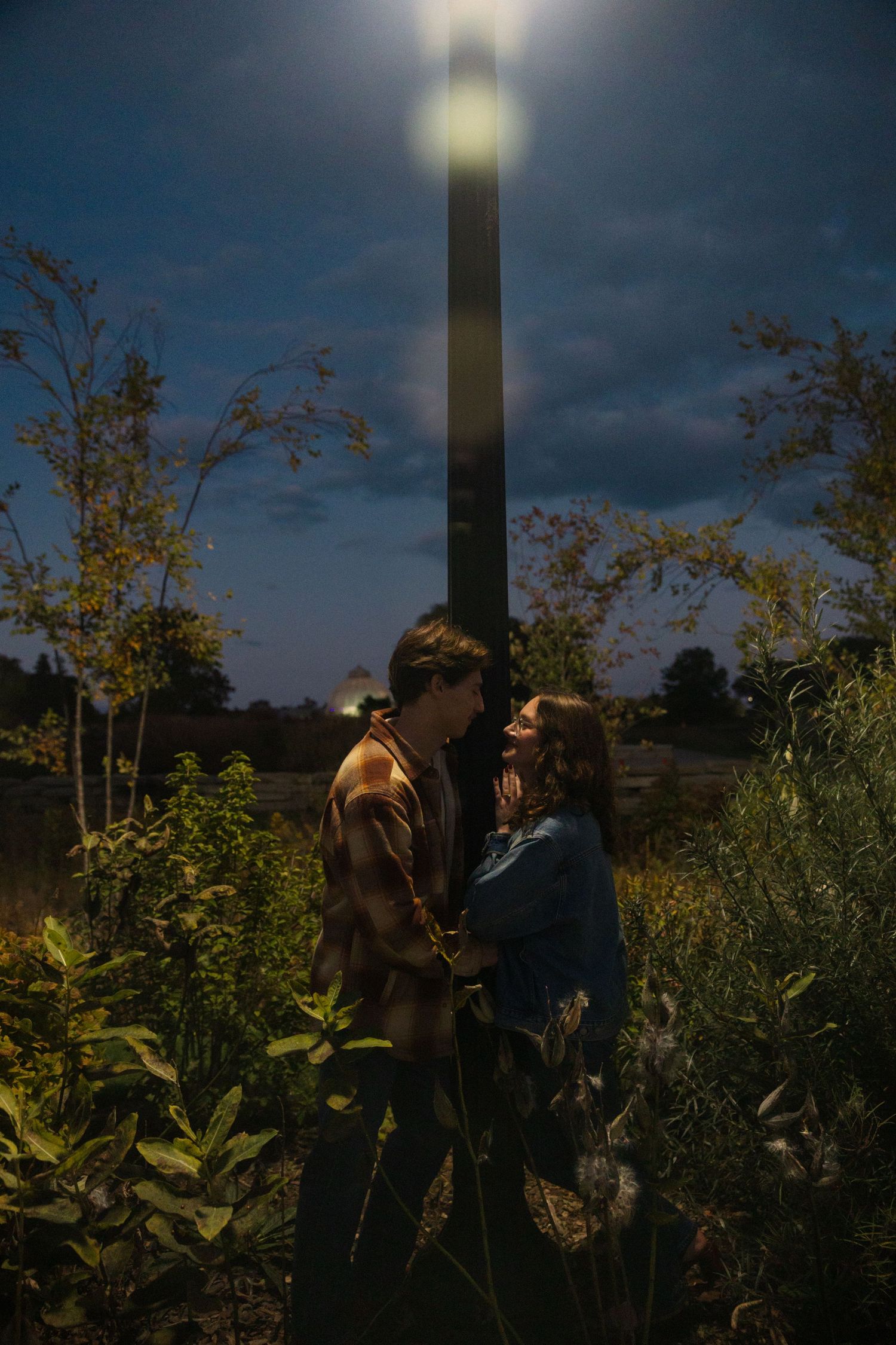 Couple embracing romantically in a lush garden under a glowing street lamp at dusk.
