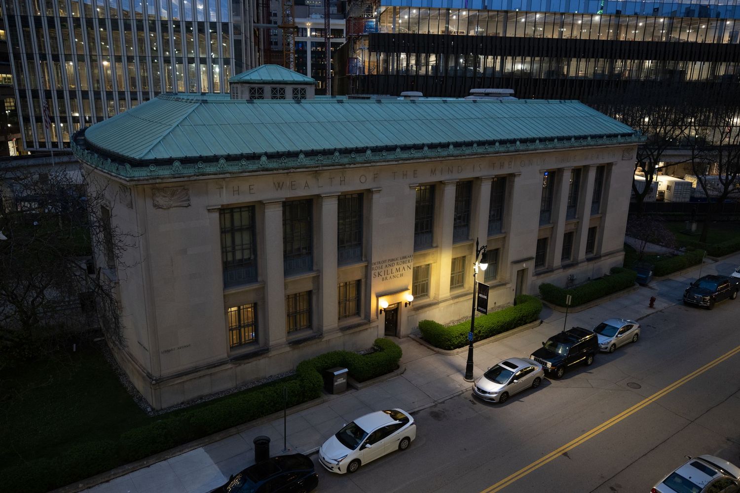 Aerial view of a neoclassical stone building with green copper roof, surrounded by parked cars on a city street at dusk.