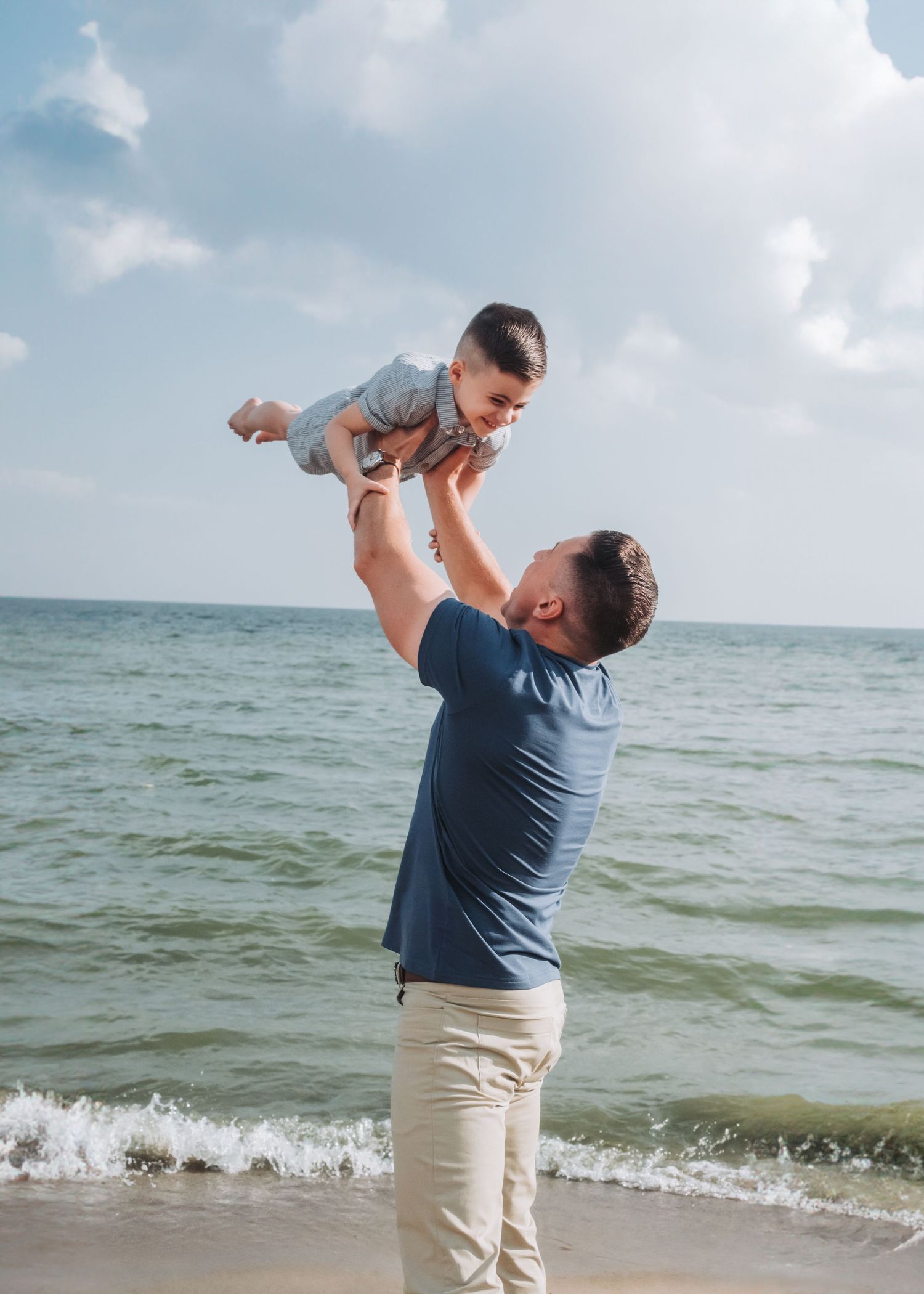 A father joyfully lifts his baby into the air on a sunny beach with ocean waves in the background.