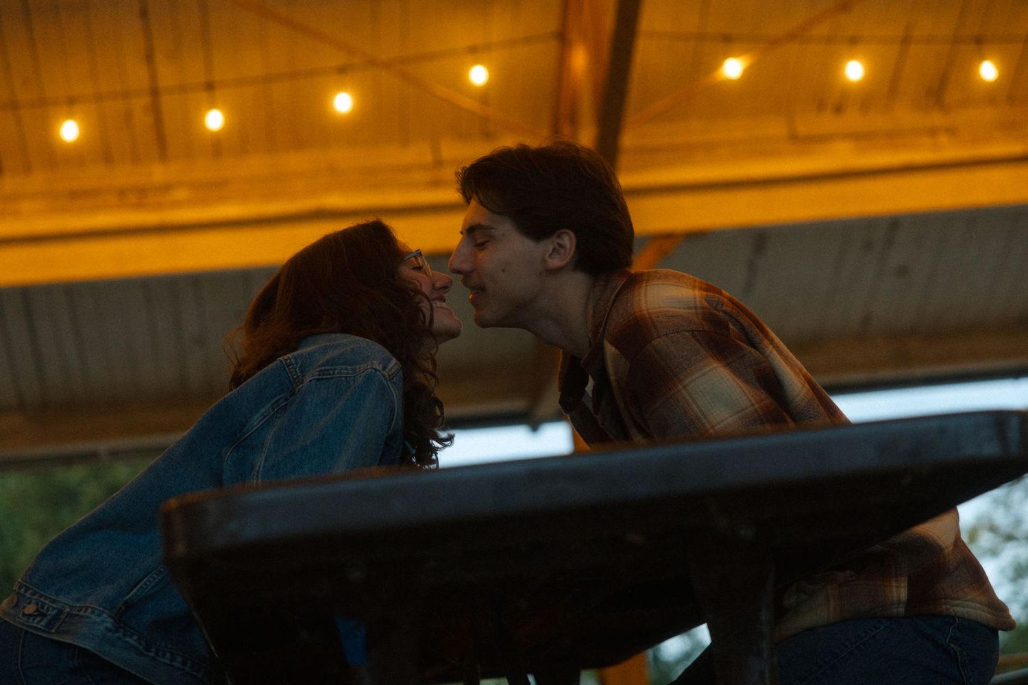 Romantic couple sharing a kiss under warm string lights at an outdoor venue at dusk.
