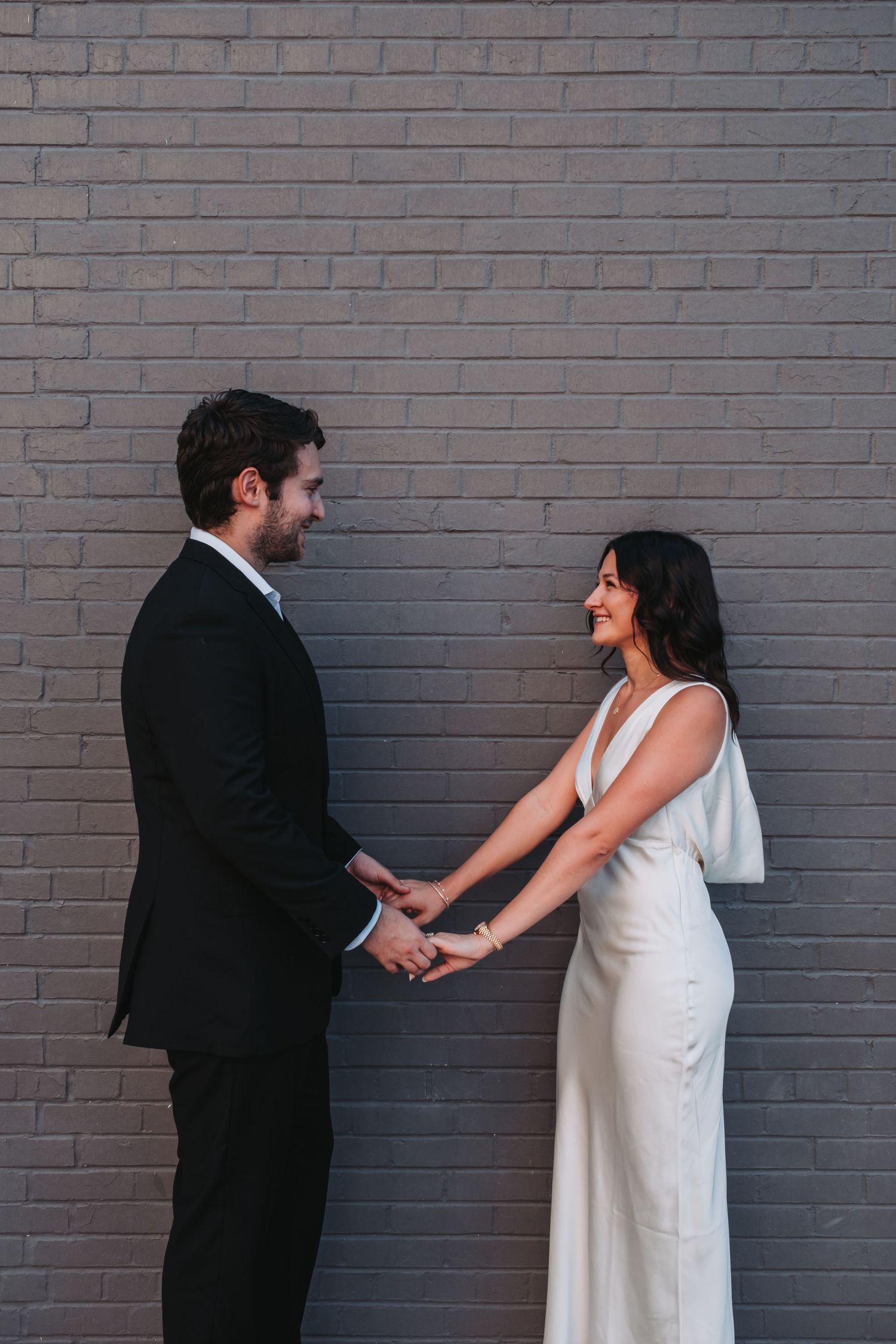 A bride and groom hold hands and smile at each other in front of a gray brick wall.