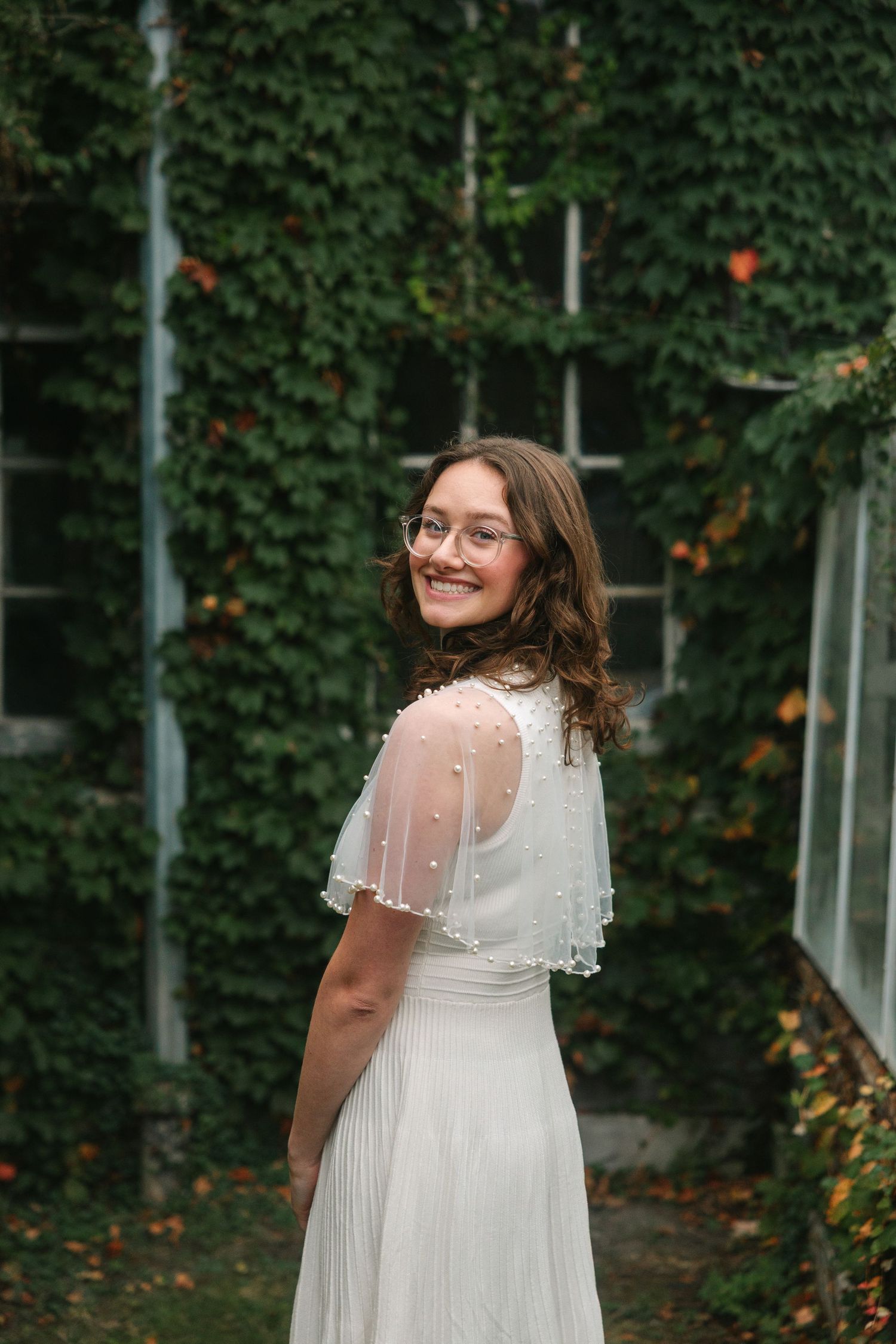 A person in a white lace dress poses in front of a green hedge wall with orange flowers.