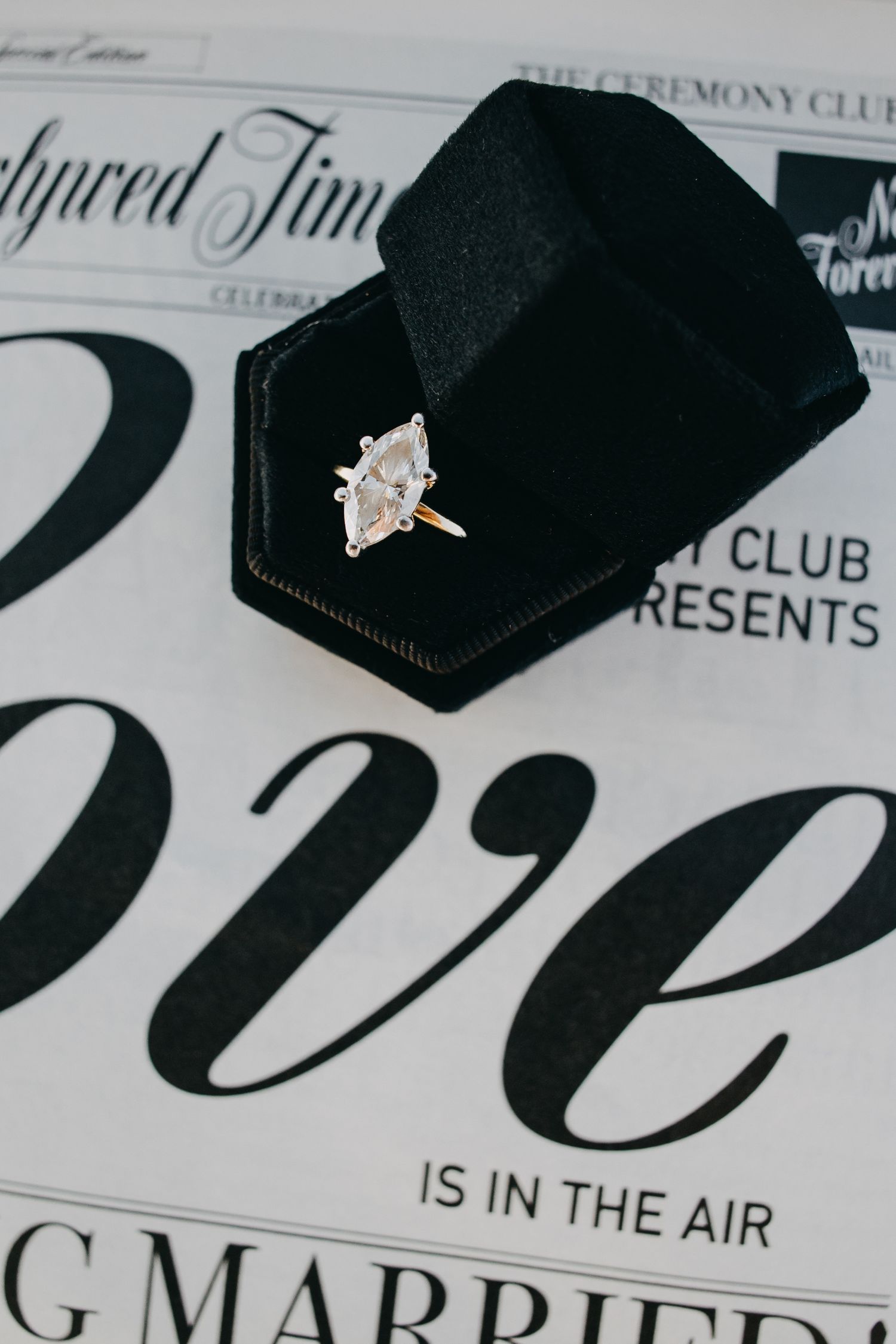 Marquise diamond engagement ring in gold setting displayed in black velvet box on wedding newspaper backdrop.