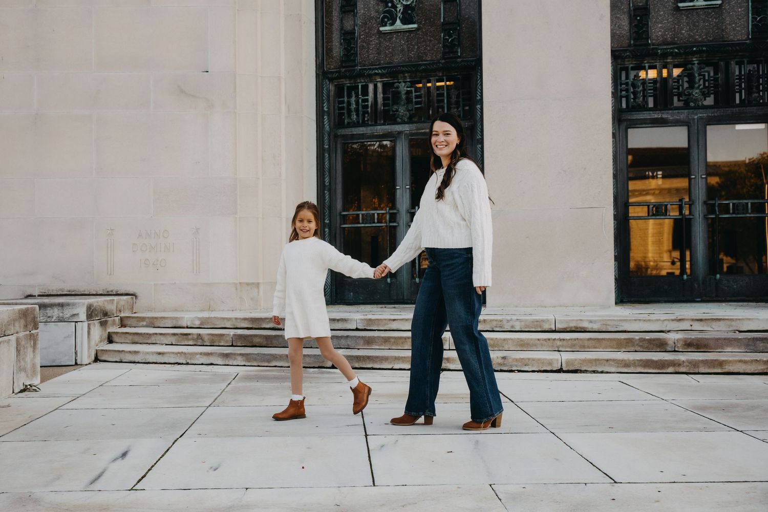 A mother and daughter hold hands in white outfits on city steps, smiling at the camera in an urban setting.