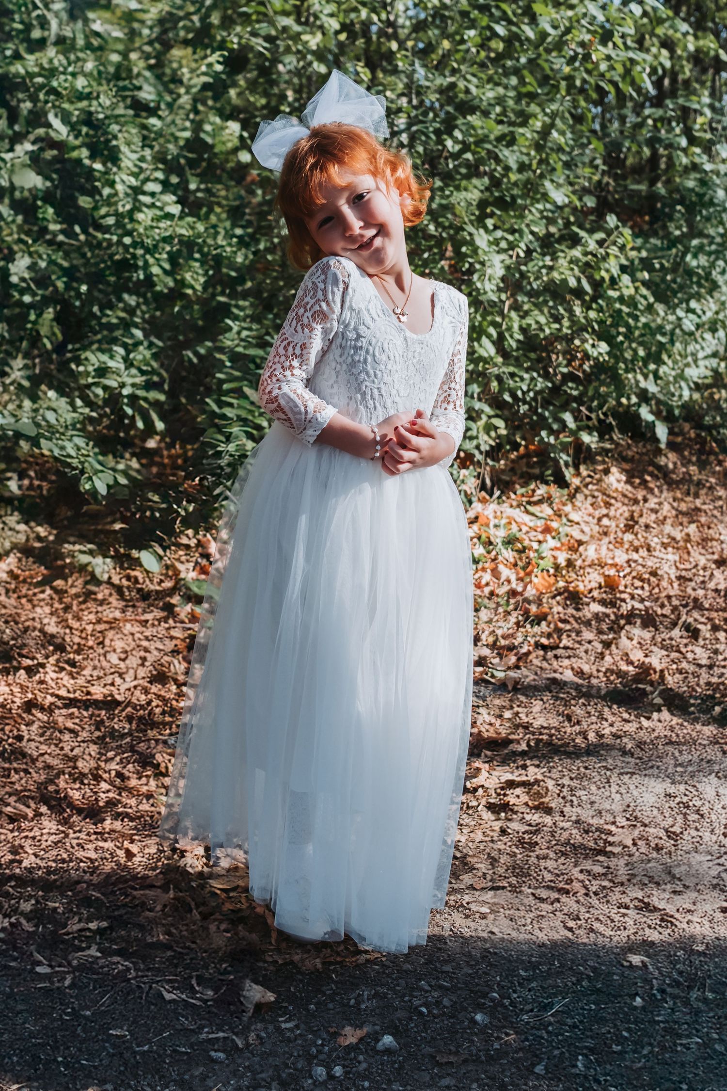 Young redhead girl smiling in white lace tulle flower girl dress with bow, standing outdoors among green bushes.