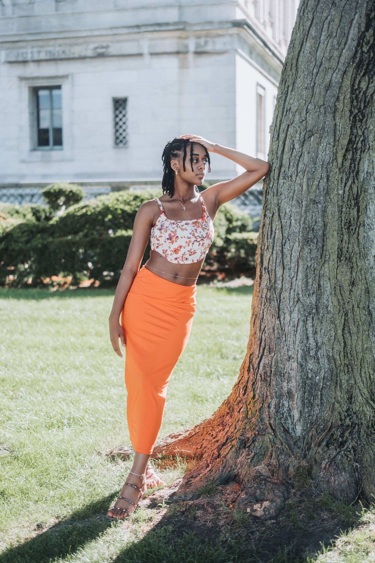 Young Black woman in orange midi skirt and floral crop top poses by a large tree on a sunny day.
