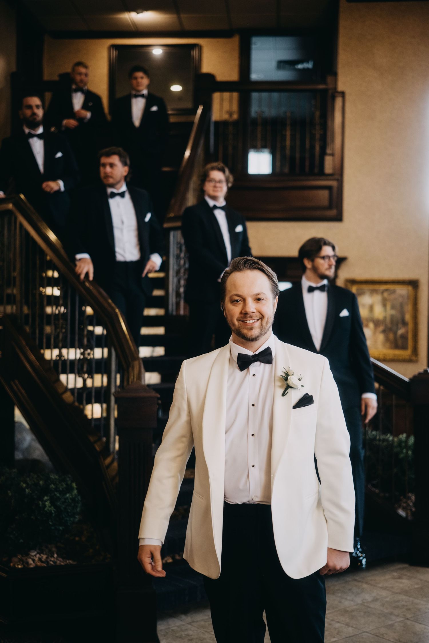 Groom in white tuxedo jacket with boutonniere stands before groomsmen in black tuxedos on elegant staircase.