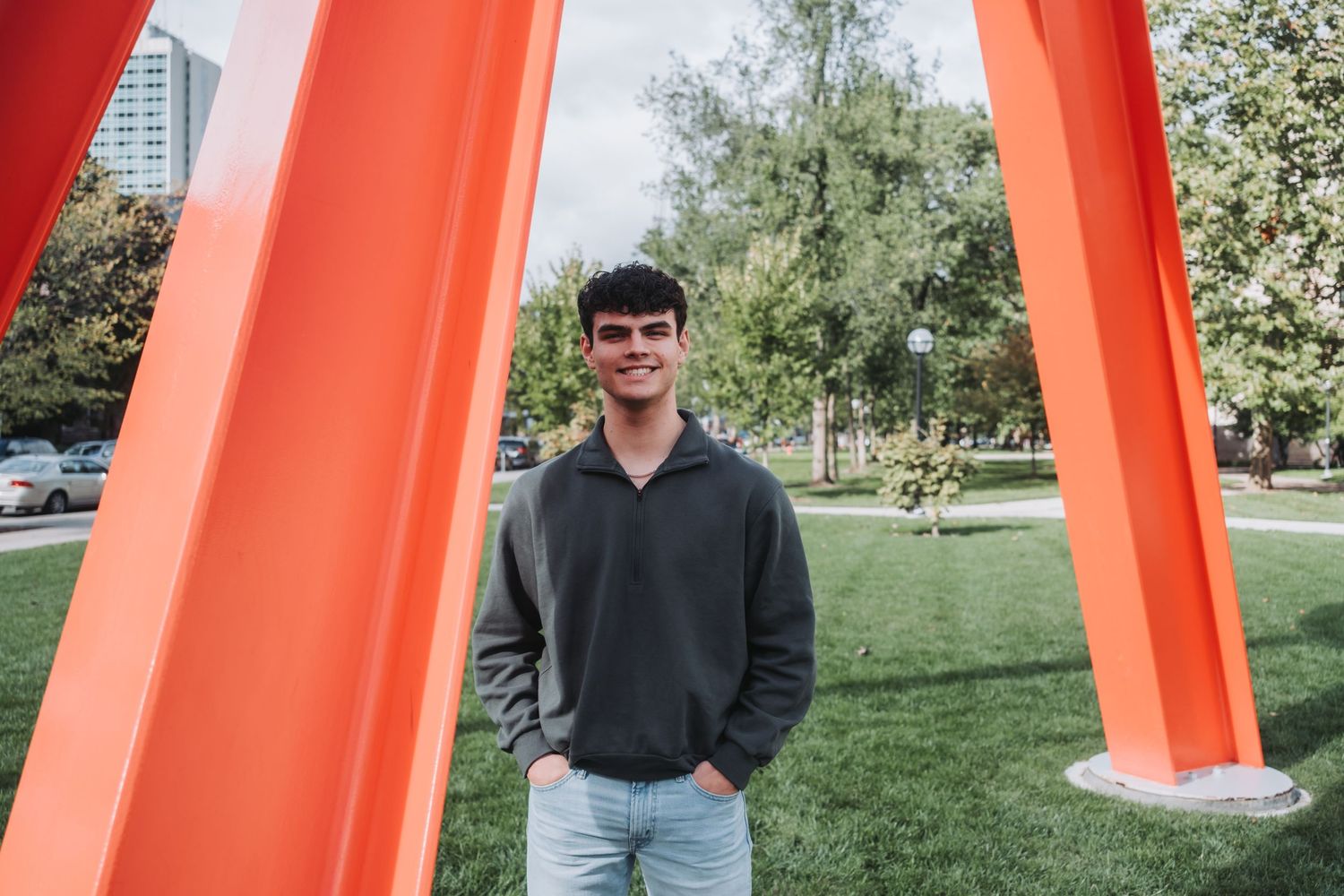 Young man in gray hoodie smiling outdoors near large orange sculpture in a green park setting.
