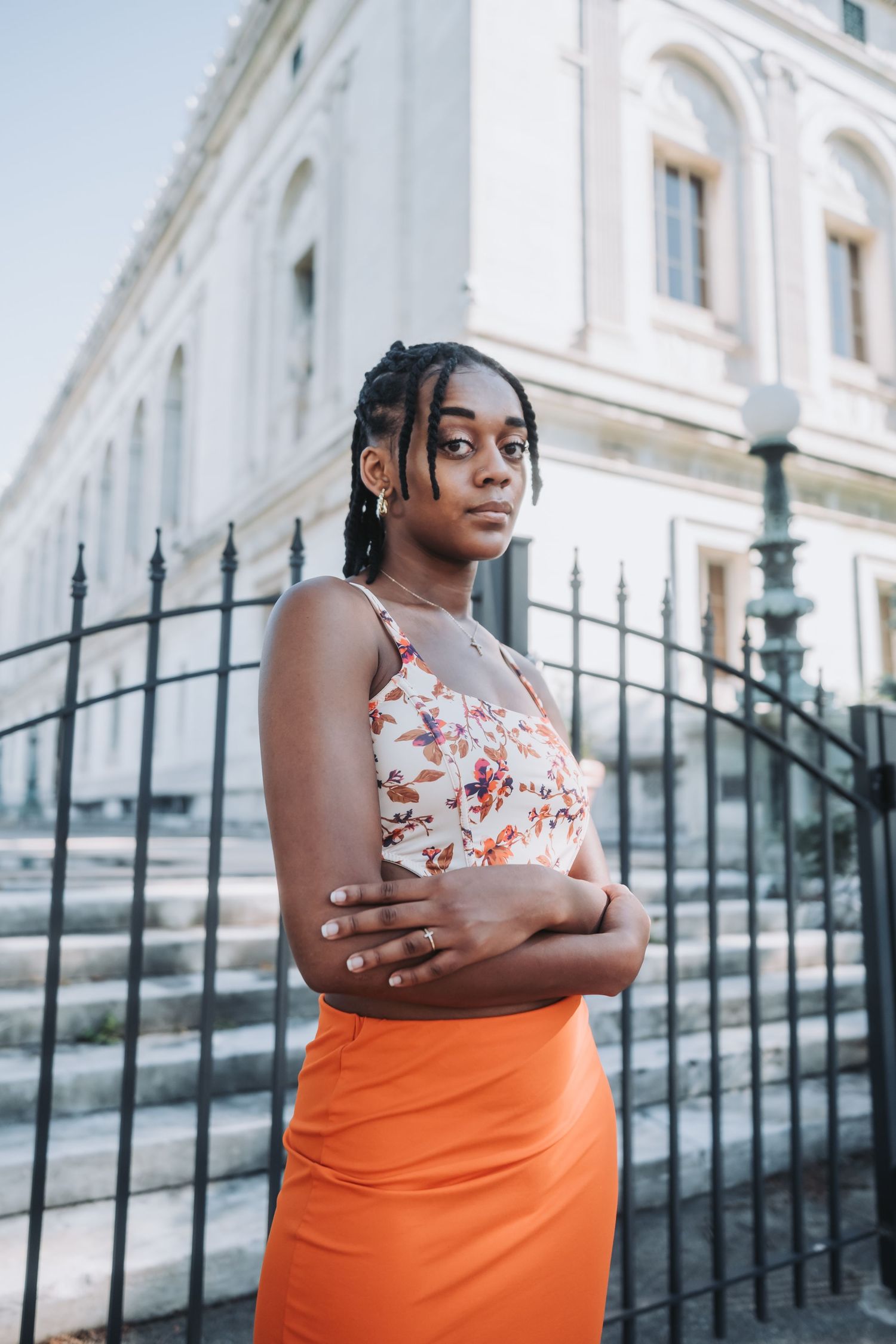 Young Black woman in floral top and orange skirt stands confidently with arms crossed before a historic building.