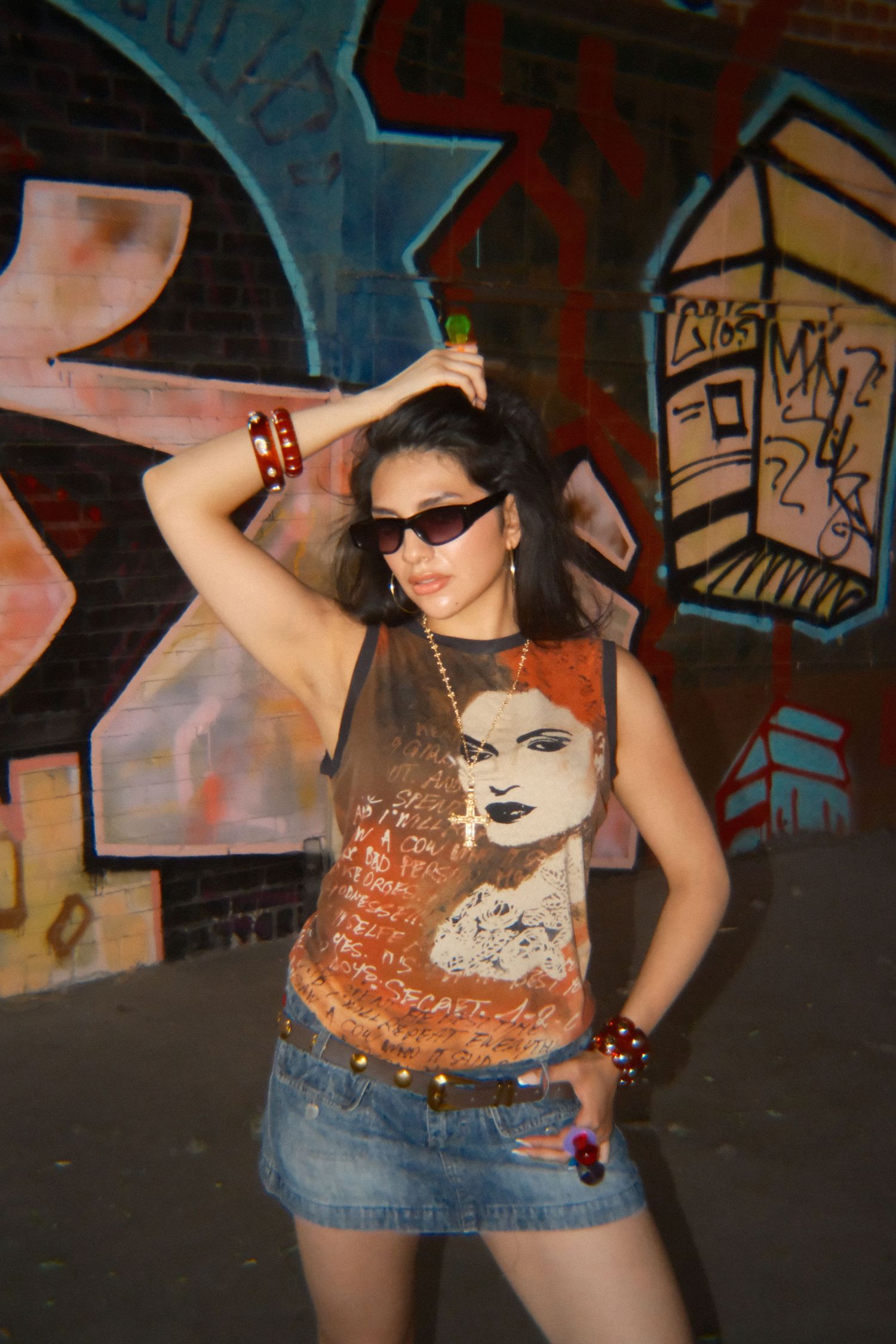 Young woman in graphic tee, denim skirt, and sunglasses posing in front of colorful graffiti wall.