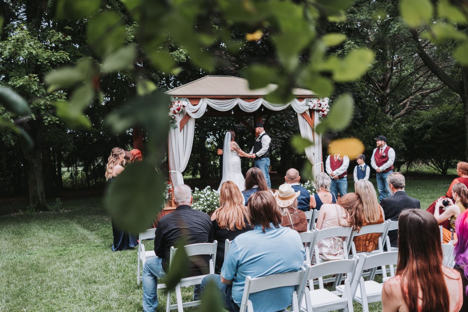 Outdoor wedding ceremony under white gazebo with guests seated on white chairs.