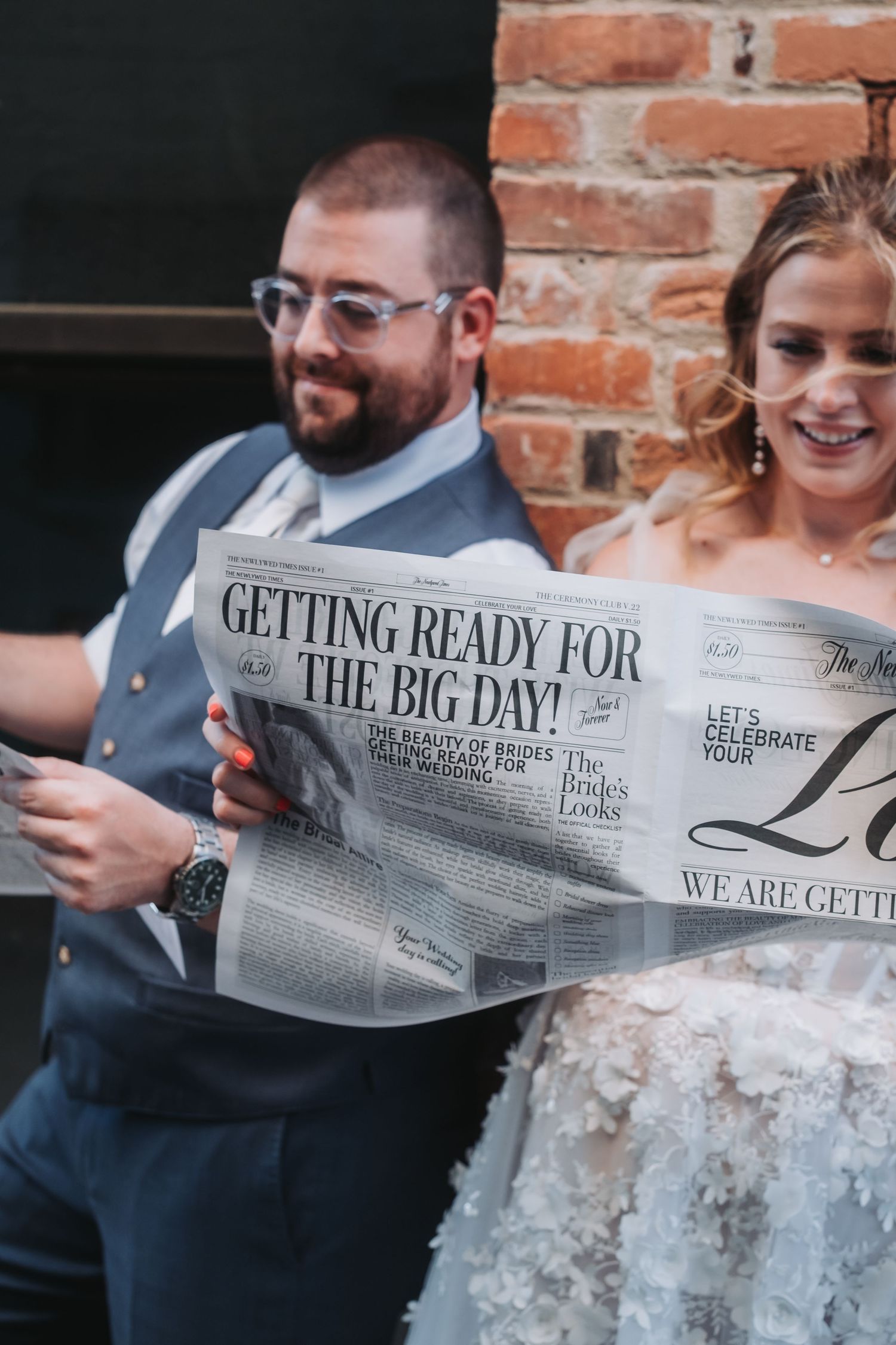 Couple reading custom wedding newspaper against brick wall background.