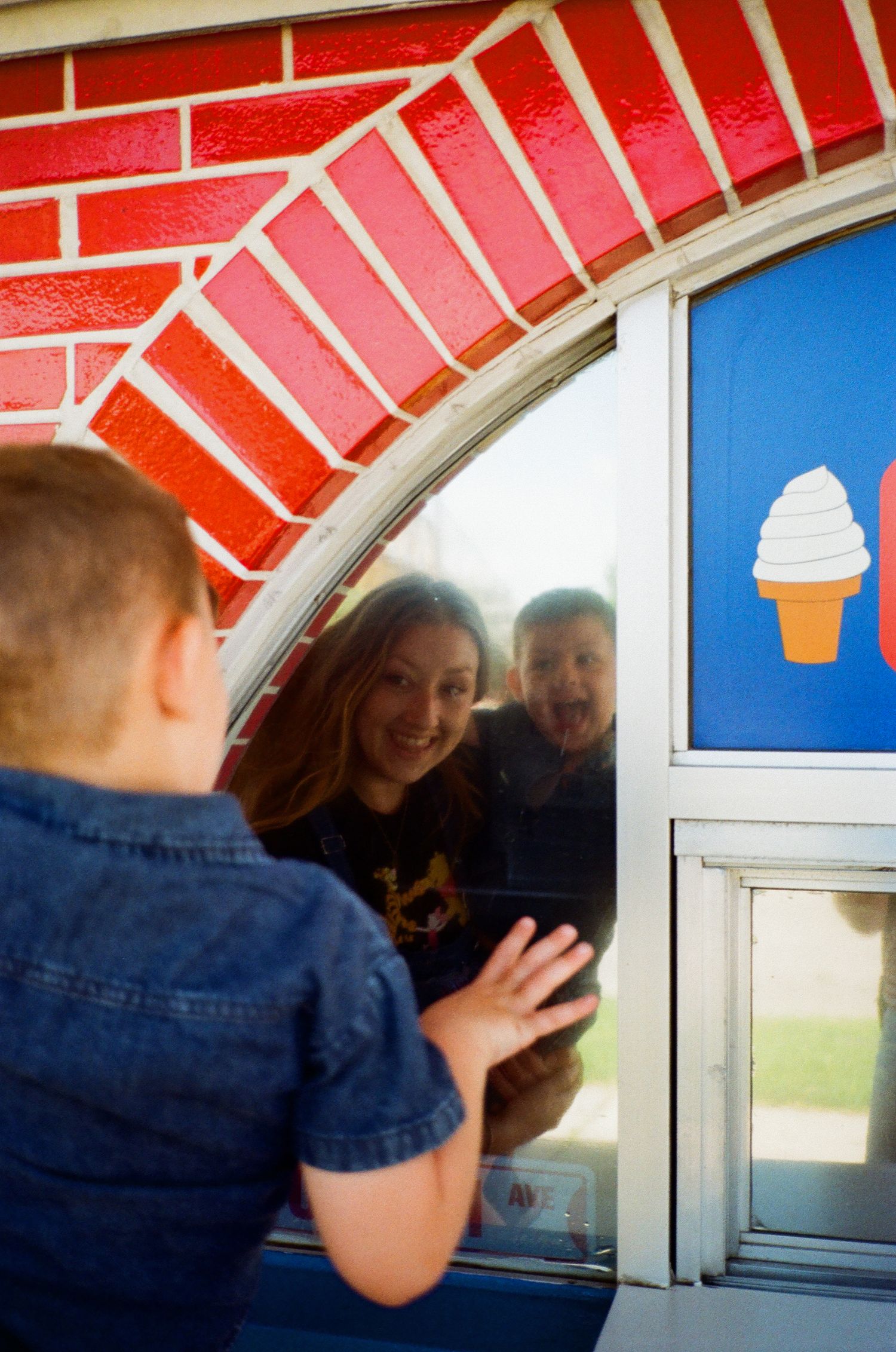 Child at ice cream shop window with smiling staff visible through glass, soft serve cone sign on blue door.