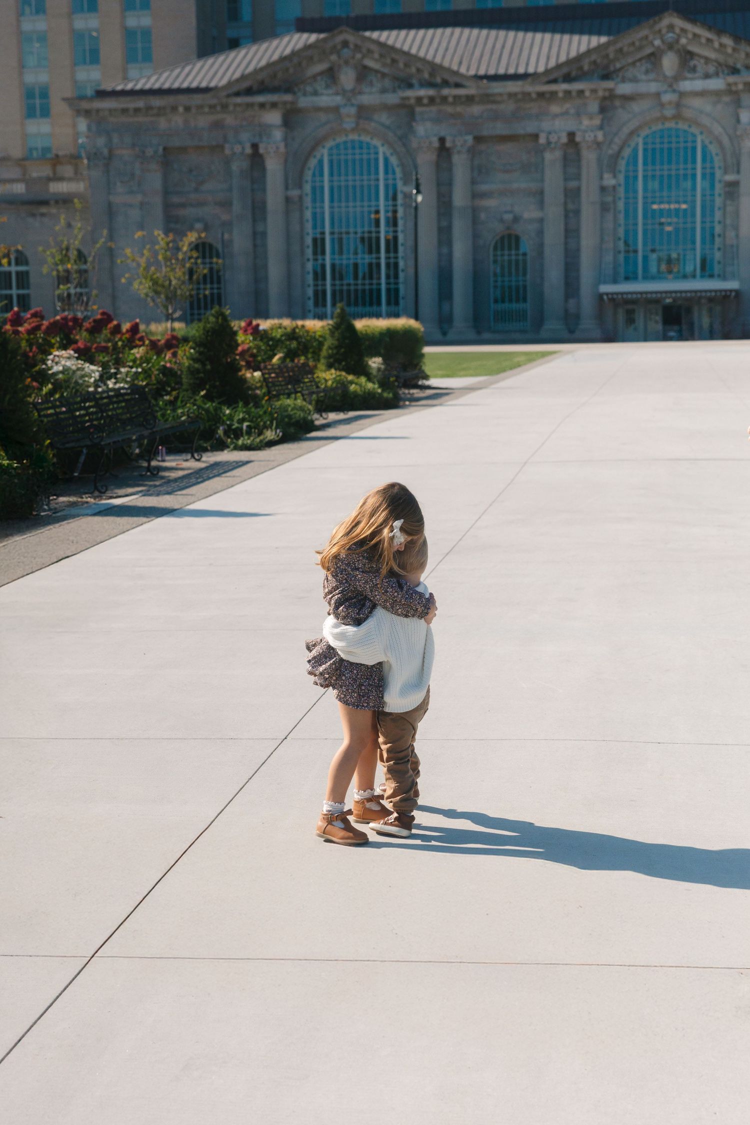 Two children hugging on a sunny plaza outside a historic stone building with arched windows.