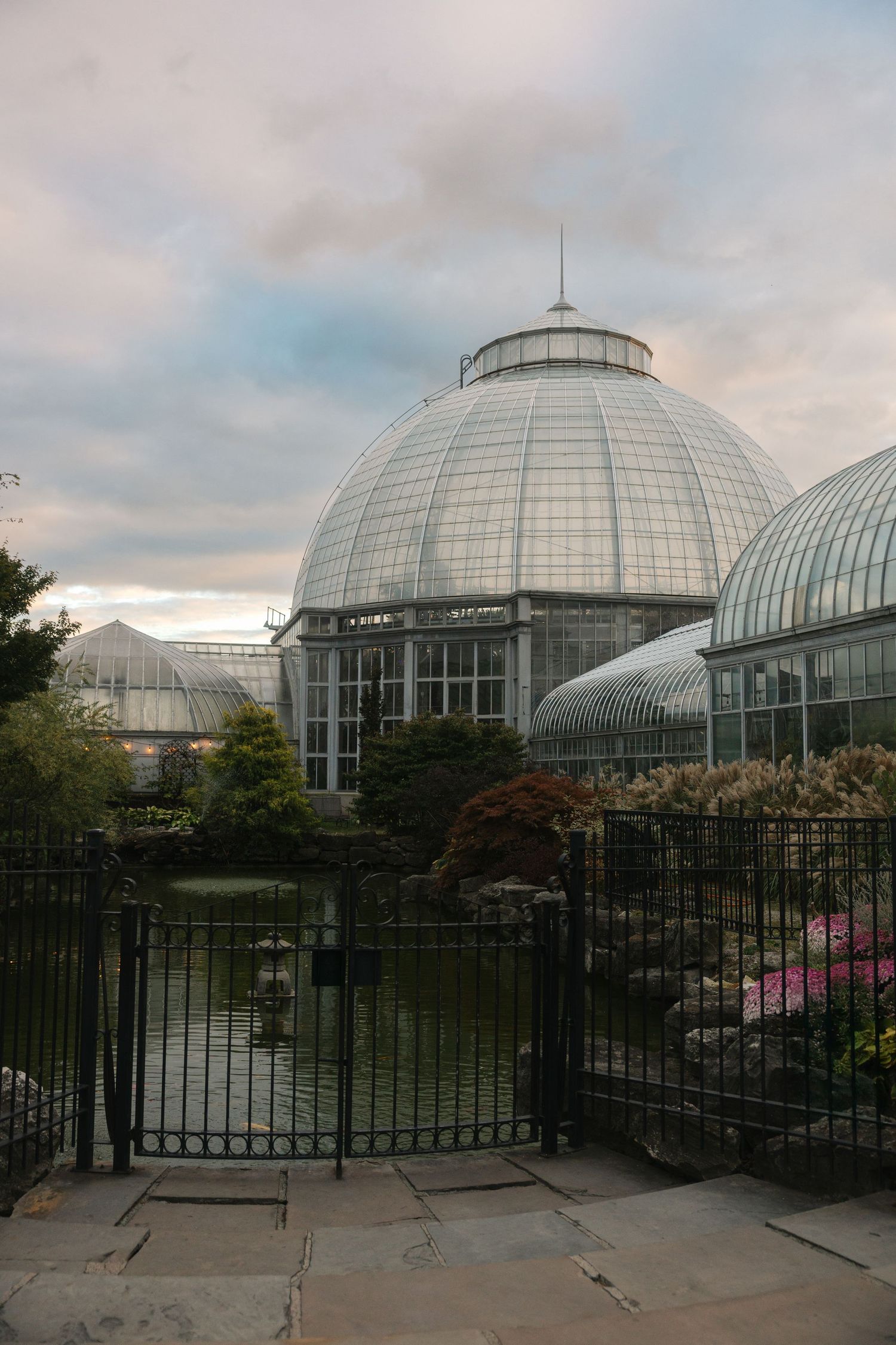Glass dome greenhouse conservatory with iron gate, pond, and colorful flowers under cloudy sky.