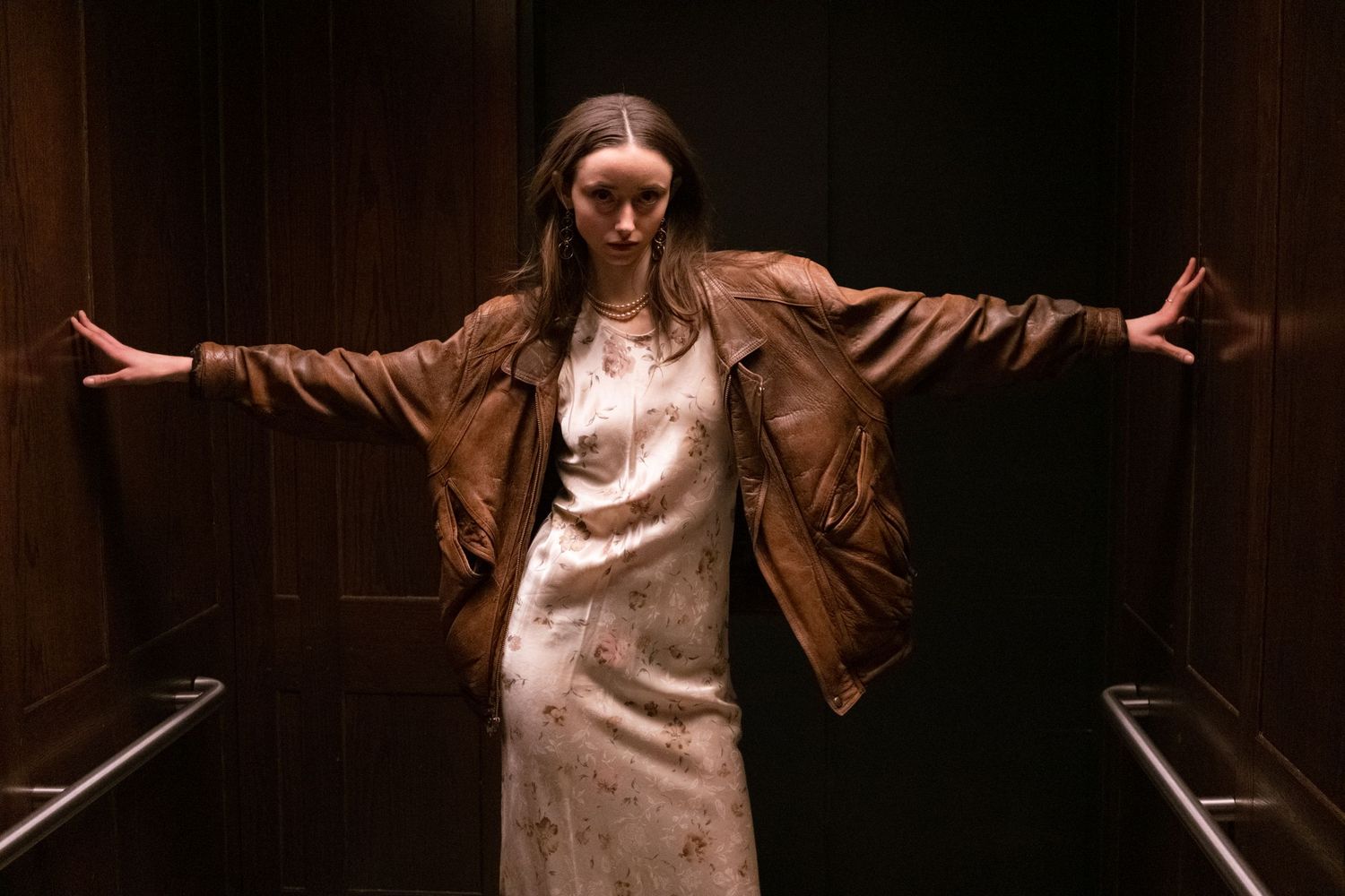A young woman in a brown leather jacket and floral dress stands with arms outstretched in a dark elevator.