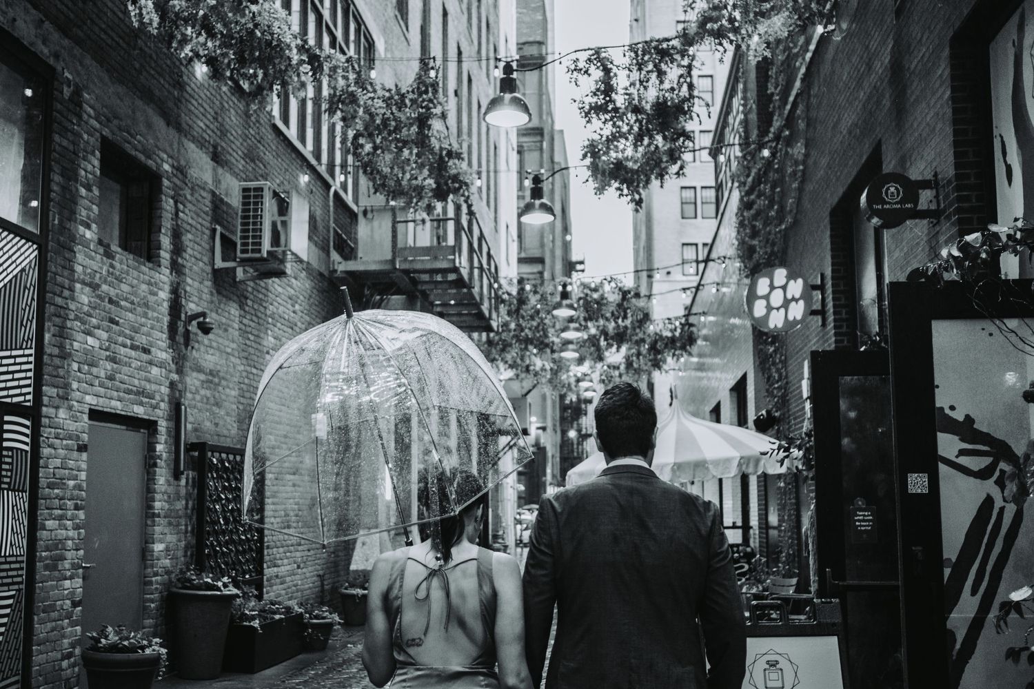 Black and white photo of a couple walking down a narrow alley sharing a clear umbrella on a rainy day.